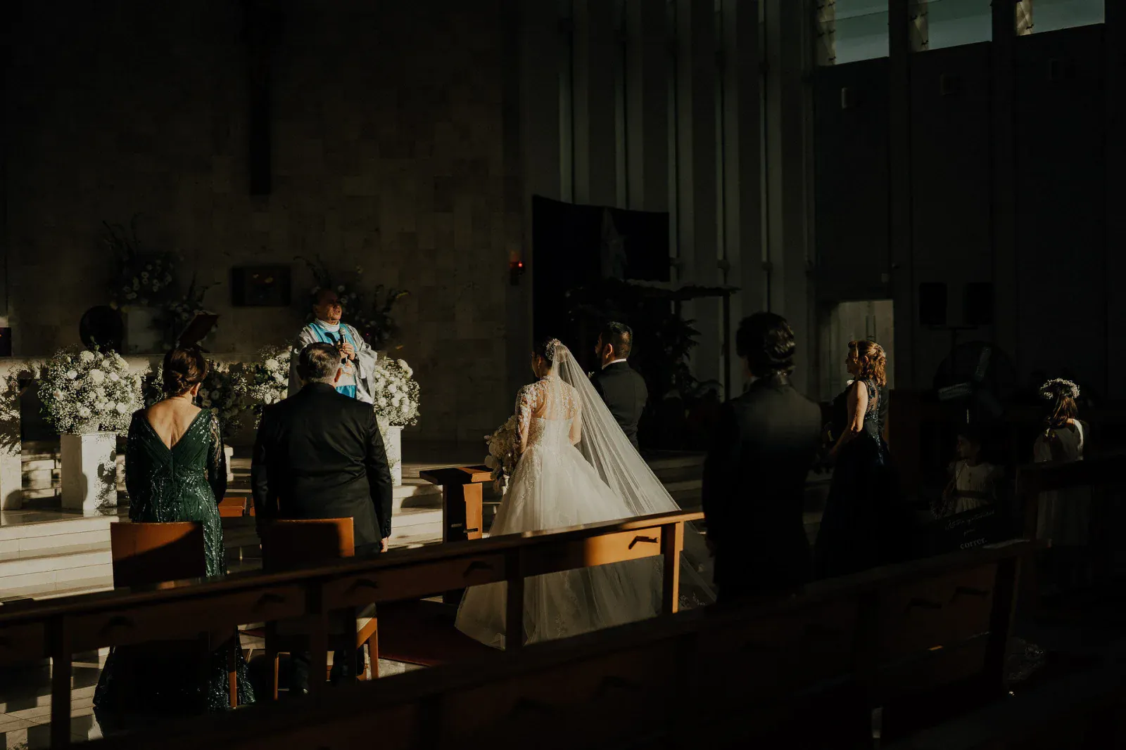Una ceremonia nupcial en una iglesia con poca luz, con una novia con un vestido vaporoso y velo, frente a un sacerdote en un altar con arreglos florales. Los invitados observan solemnemente.