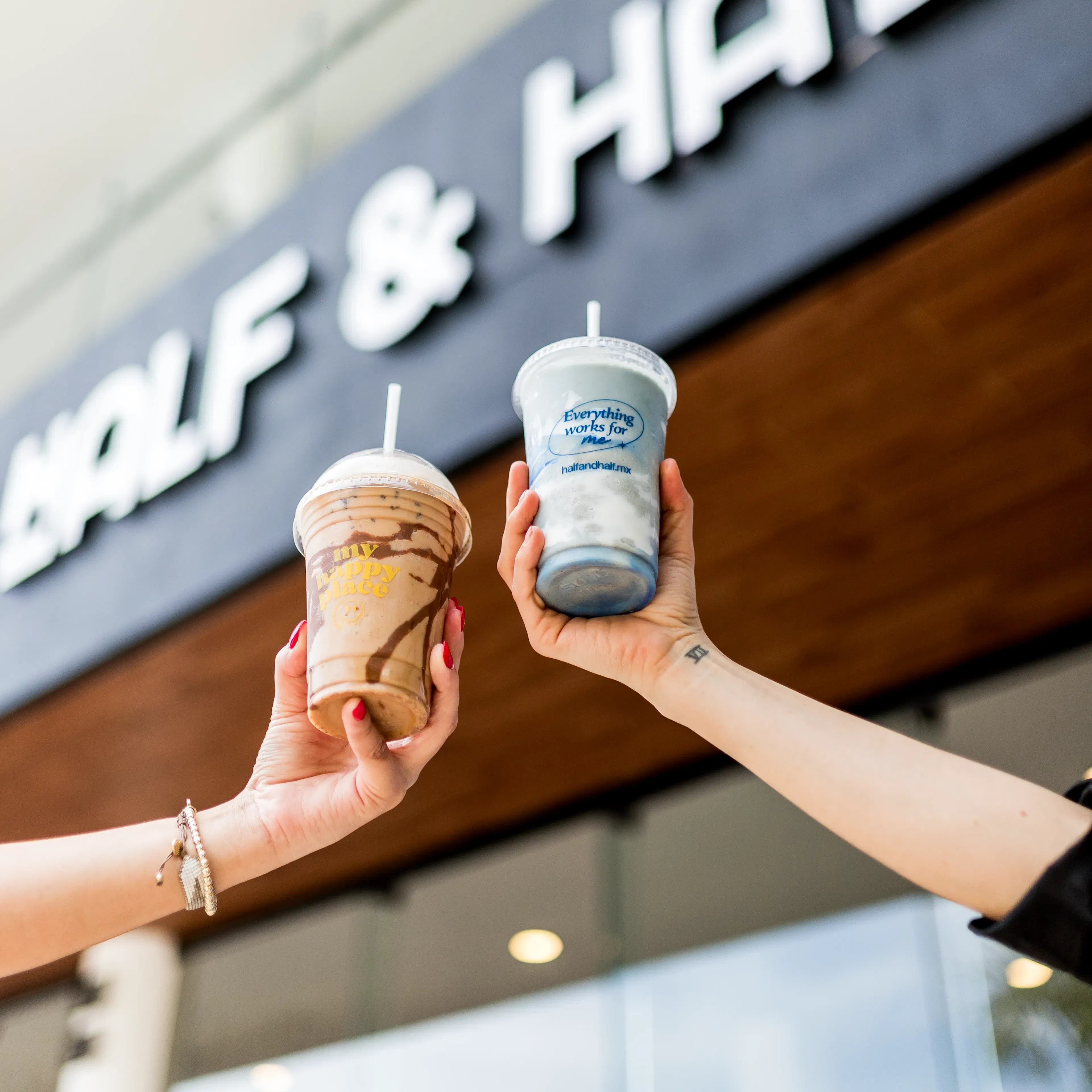 Dos manos sostienen bebidas heladas con pajitas frente a un cartel que dice "Half & Half". La escena transmite una atmósfera alegre y refrescante.