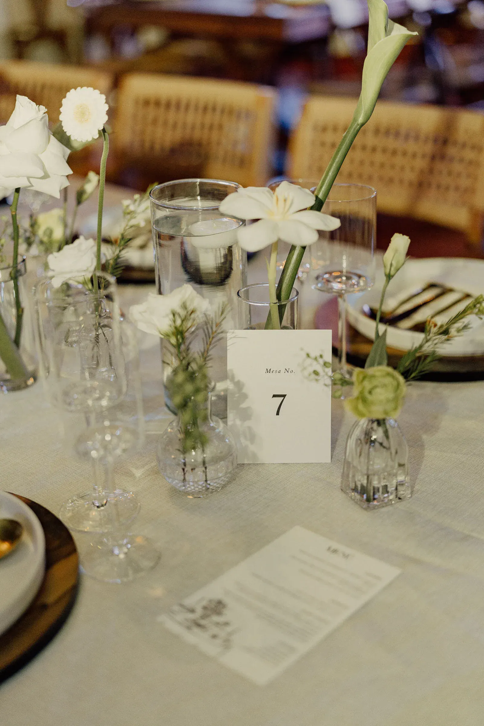 Elegante mesa con jarrones de cristal con flores blancas, la séptima mesa y un menú sobre un mantel blanco. El ambiente es sereno y refinado.