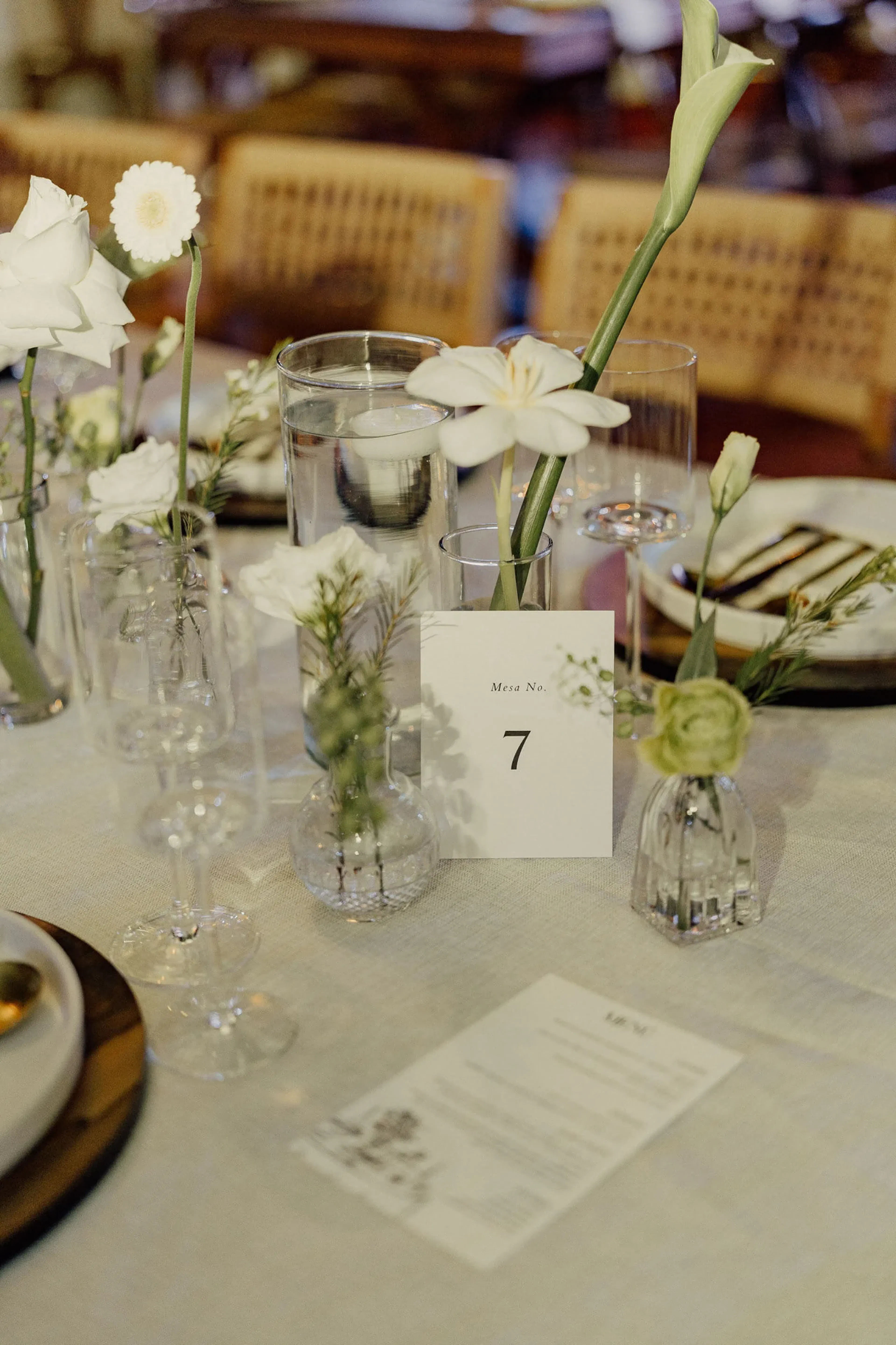 Elegante mesa con jarrones de cristal con flores blancas, la séptima mesa y un menú sobre un mantel blanco. El ambiente es sereno y refinado.