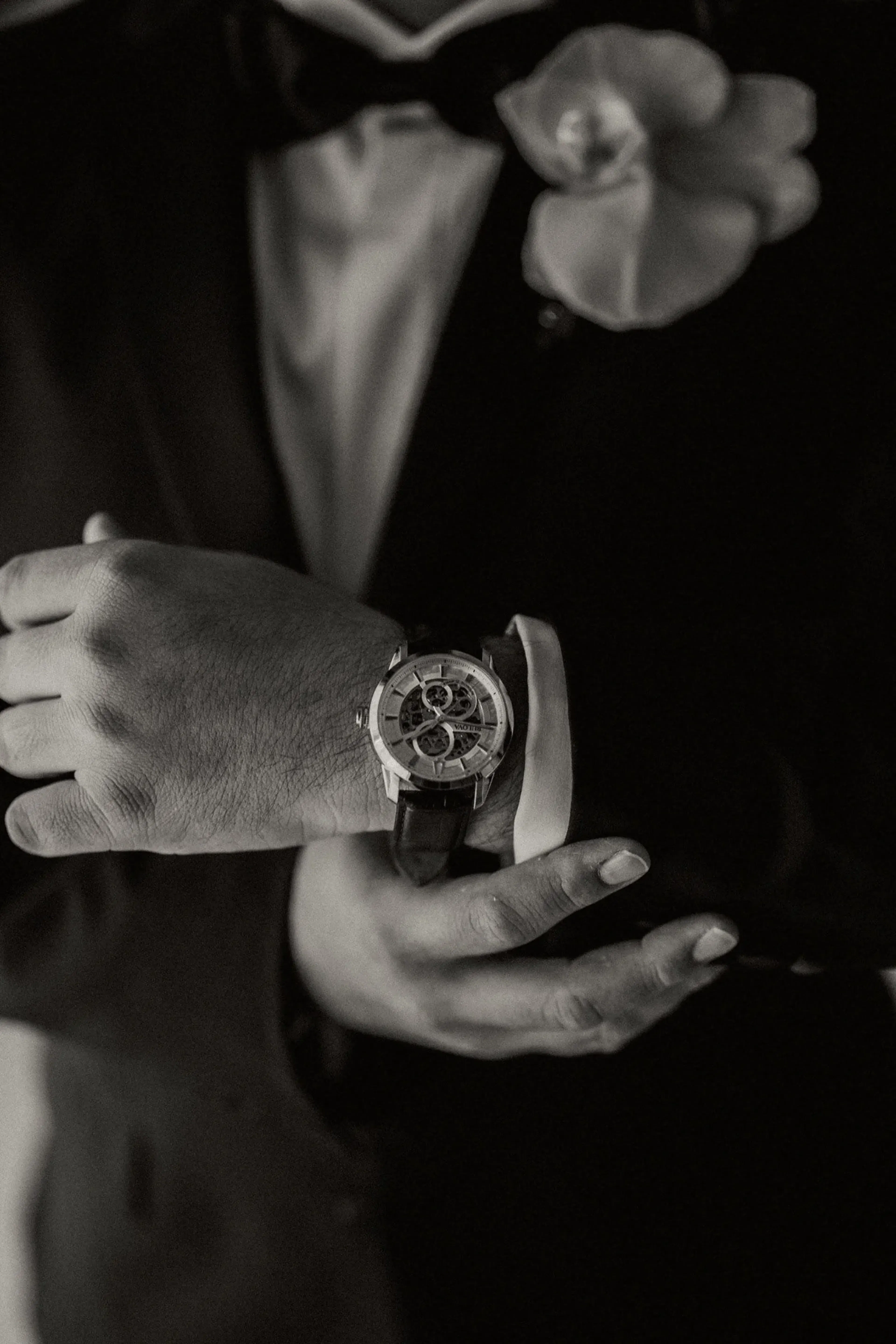 Un hombre de traje se ajusta el puño de la camisa, luciendo un intrincado reloj esqueleto. Un boutonniere decora su chaqueta, transmitiendo elegancia y sofisticación.