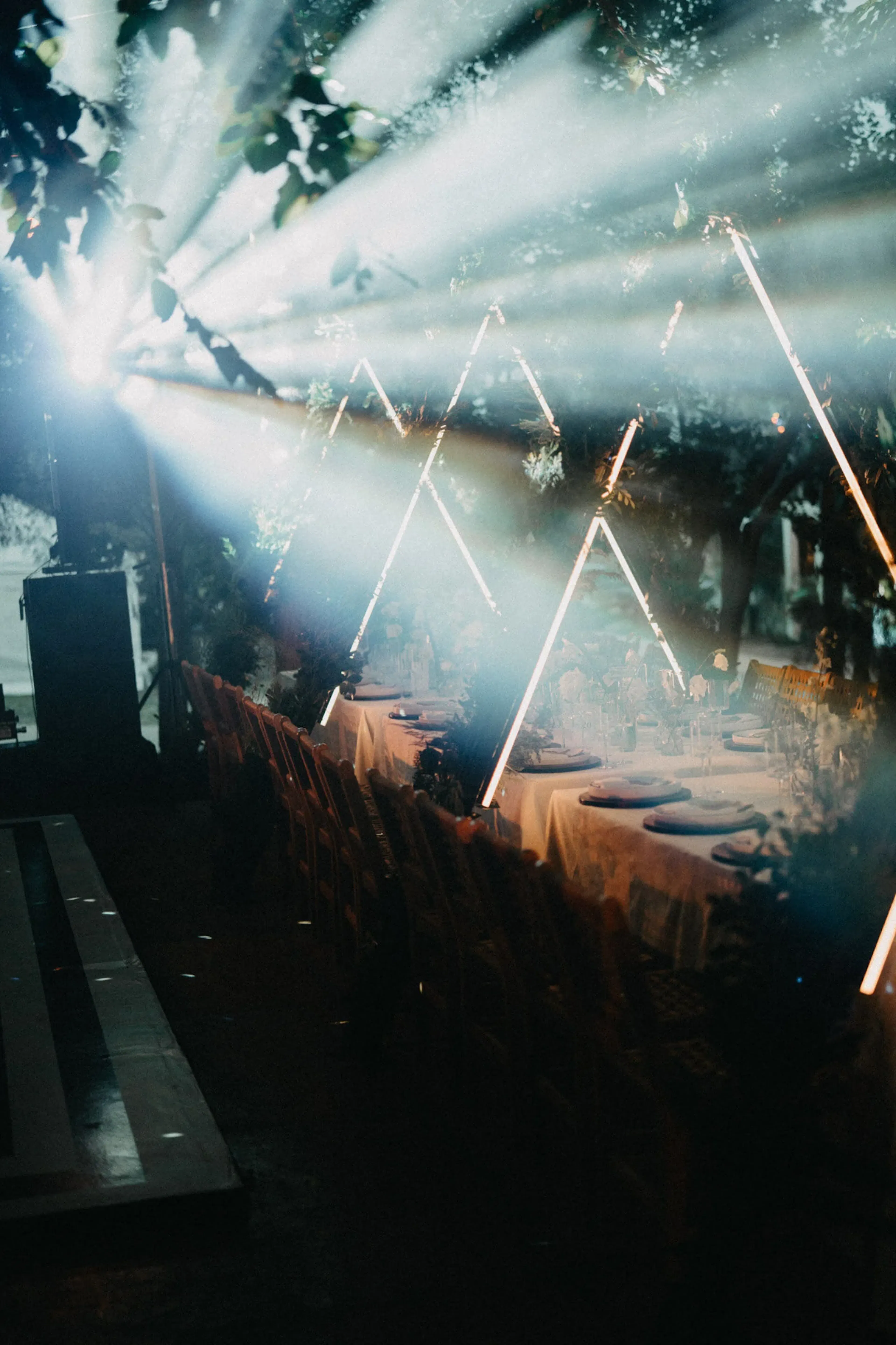 Mesa larga para cenar bajo los árboles, iluminada por una luz espectacular. Ambiente elegante con estructuras geométricas de luz.