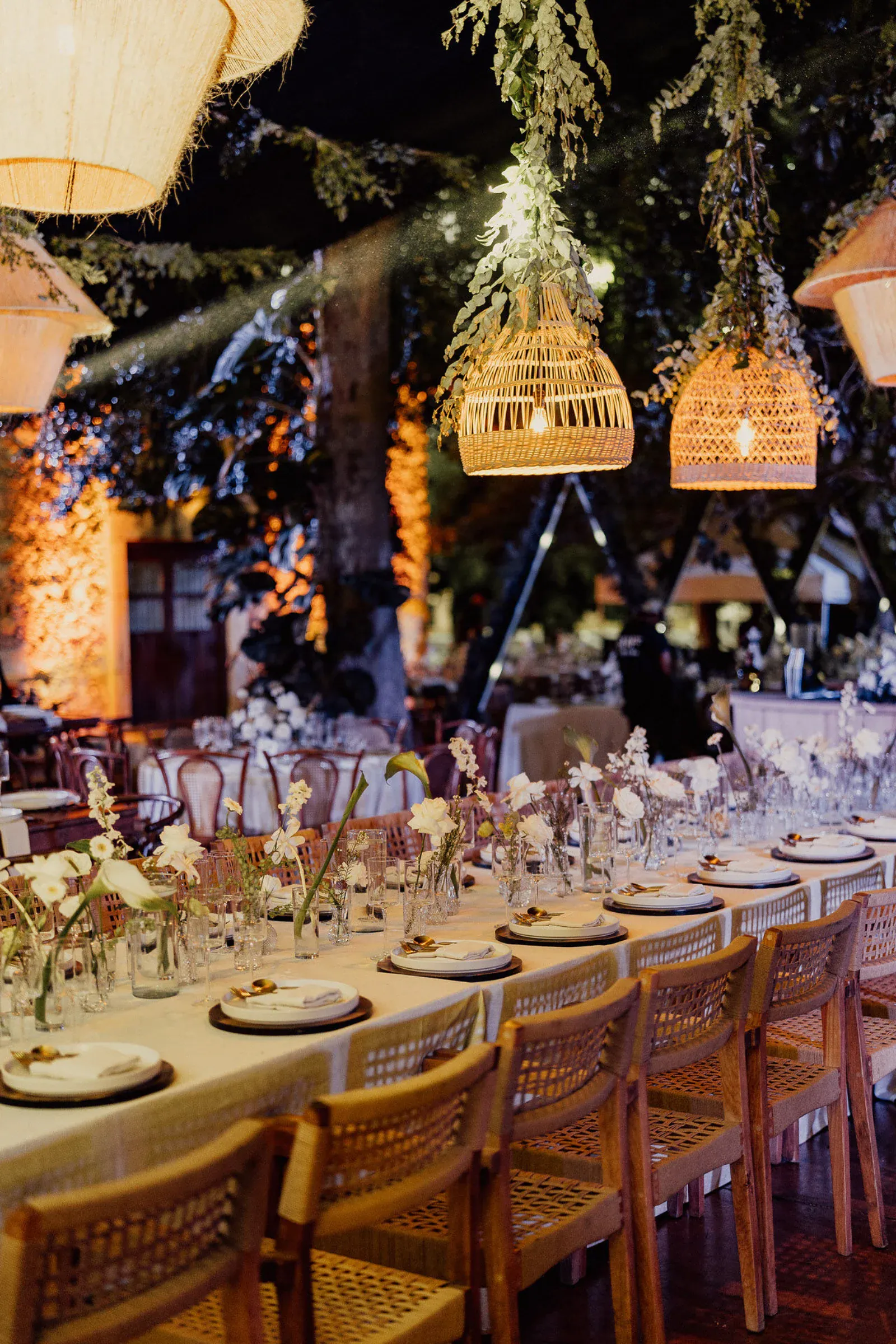 Elegante comedor al aire libre con una larga mesa iluminada con velas y adornada con flores. Lámparas colgantes tejidas y follaje cuelgan del techo, creando un ambiente cálido.
