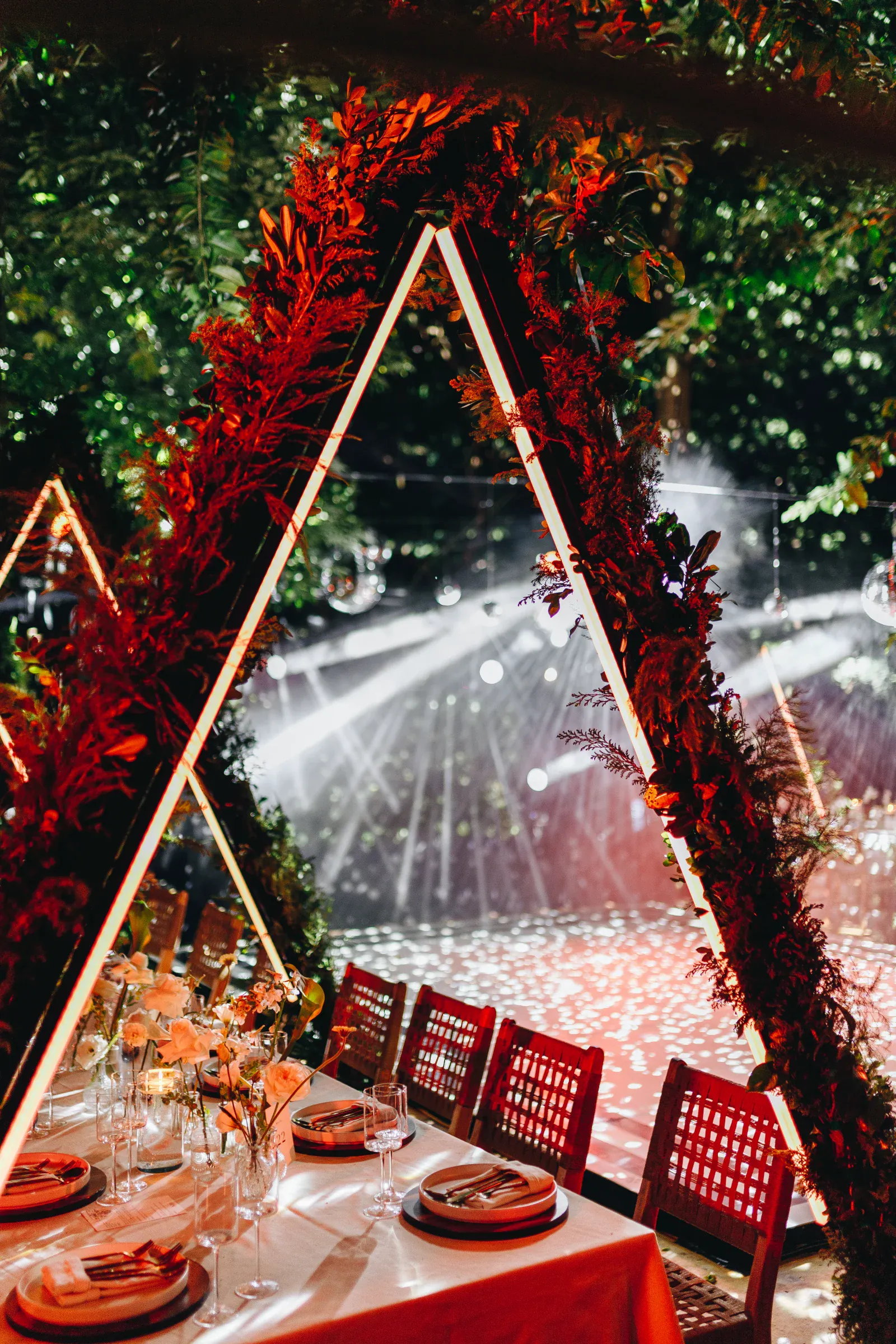 Elegante montaje para eventos con mesas bajo arcos triangulares iluminados y cubiertos de vegetación. La iluminación ambiental crea una atmósfera mágica, realzando el fondo del bosque.