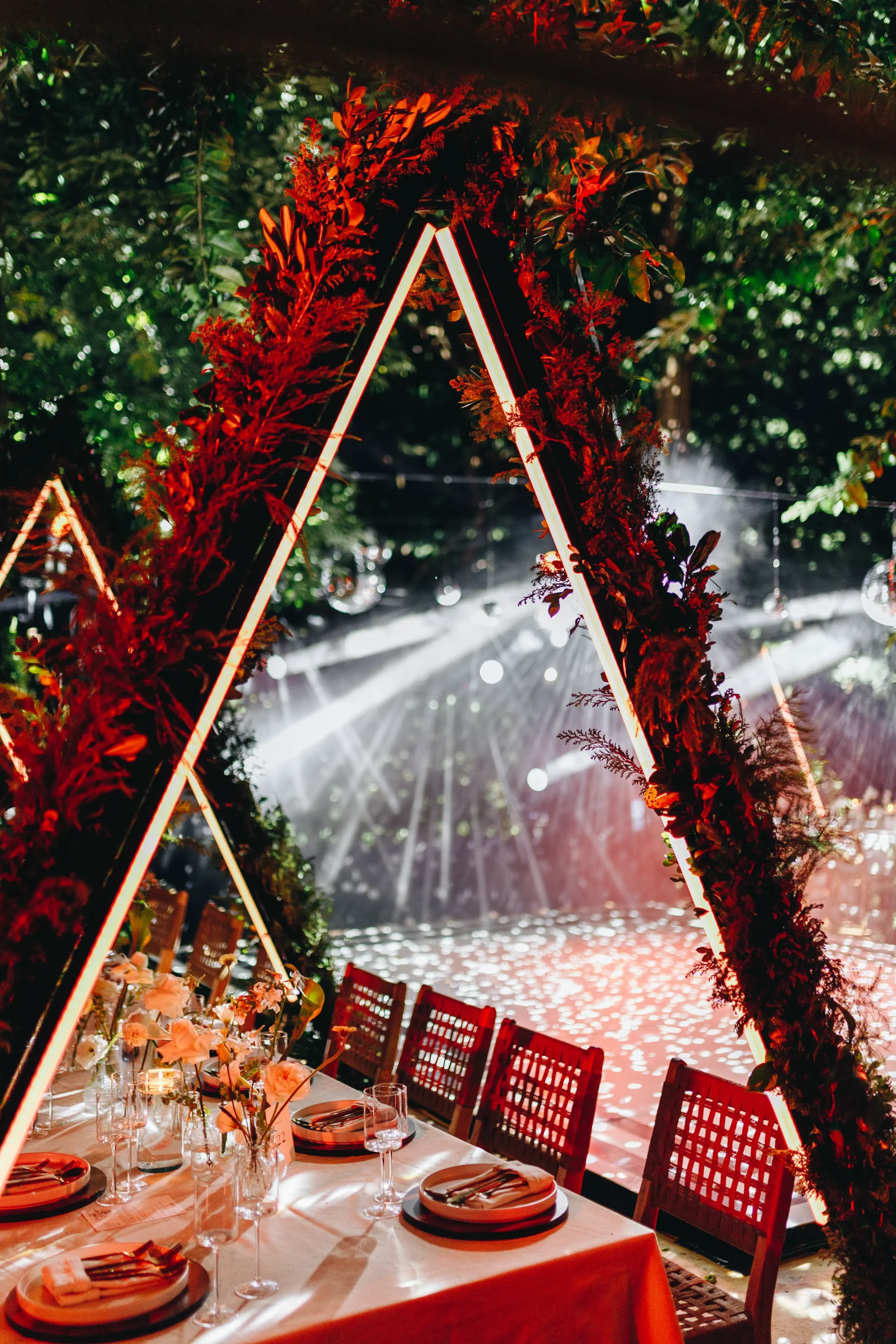 Elegante montaje para eventos con mesas bajo arcos triangulares iluminados y cubiertos de vegetación. La iluminación ambiental crea una atmósfera mágica, realzando el fondo del bosque.