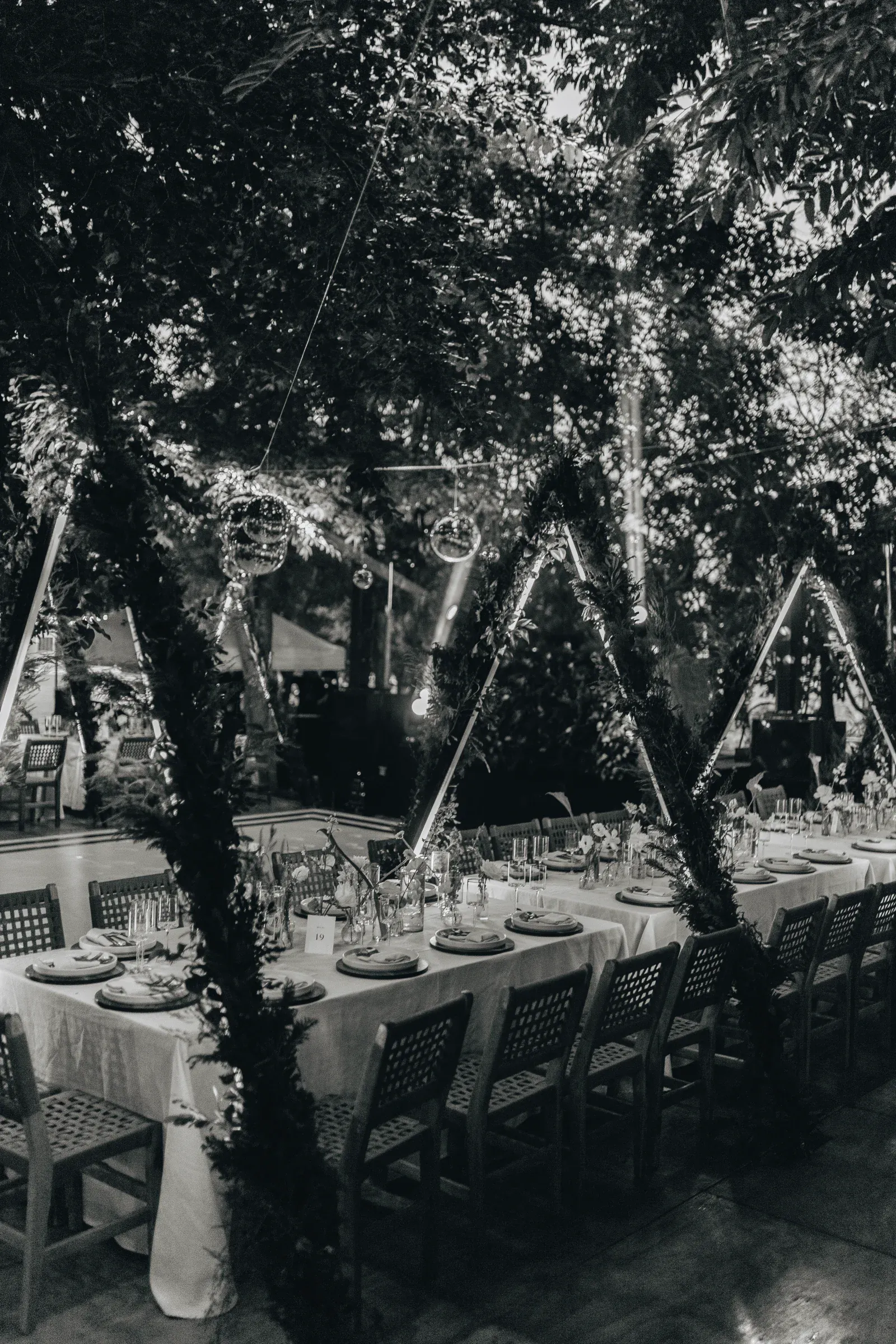 Una mesa de comedor larga y elegantemente dispuesta con mantelería blanca se encuentra al aire libre, bajo luces centelleantes y rodeada de exuberante vegetación, creando un ambiente romántico.