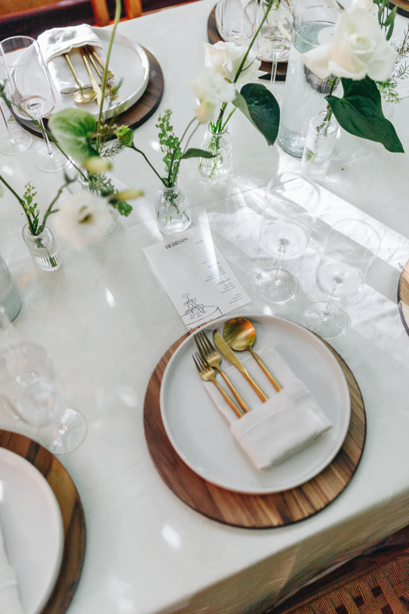 Elegante mesa con platos blancos sobre bases de madera, cubiertos dorados en servilletas blancas y delicados jarrones dispersos con rosas blancas y vegetación.
