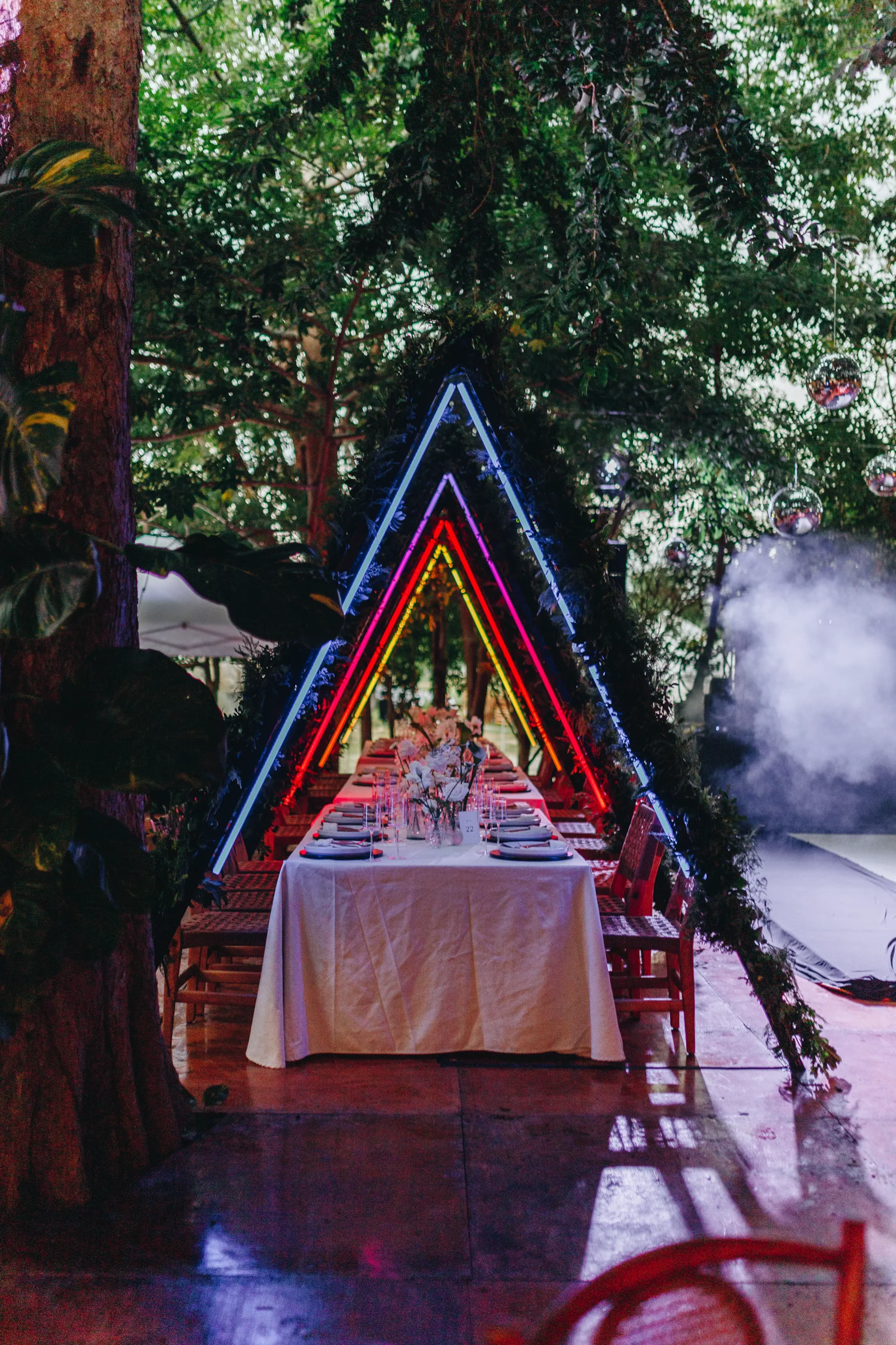 Una larga mesa de comedor bajo arcos triangulares de neón en un bosque. Las luces de colores, el humo y la exuberante vegetación crean una atmósfera mística y encantadora.