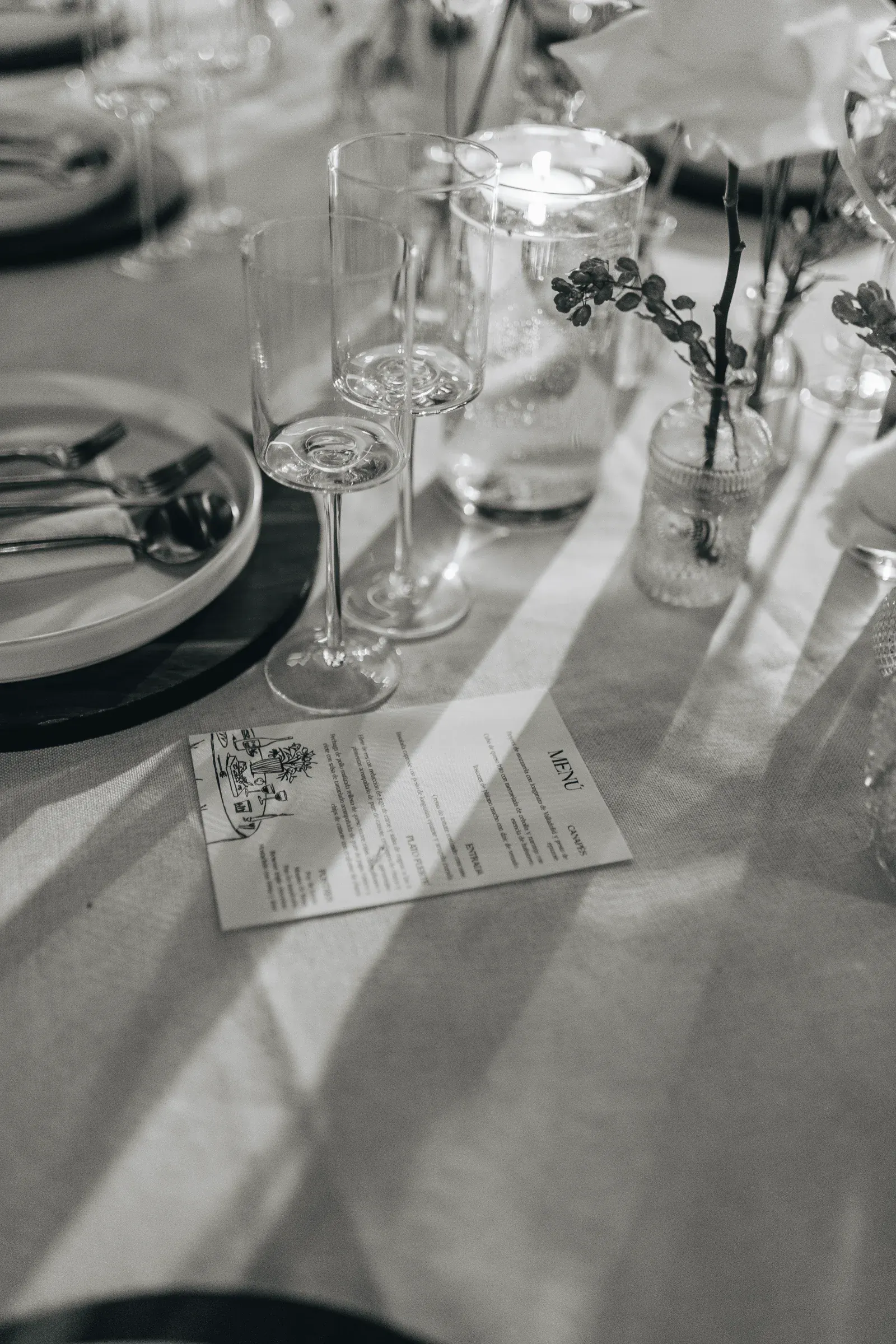 Elegante mesa con copas de vino, un plato con cubiertos y un pequeño jarrón con flores. Sobre un mantel de lino se encuentra una tarjeta de menú. Ambiente clásico y sofisticado.