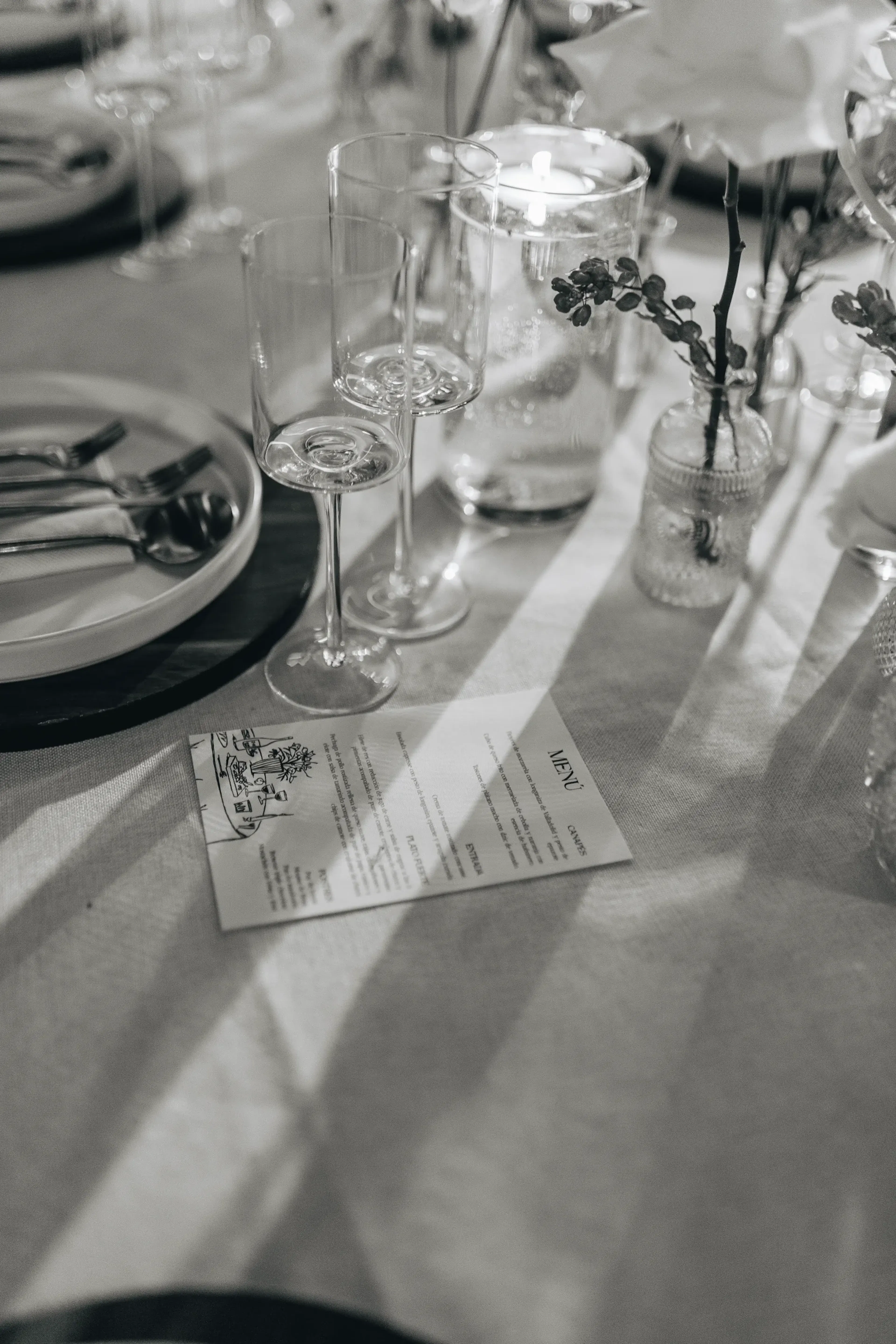 Elegante mesa con copas de vino, un plato con cubiertos y un pequeño jarrón con flores. Sobre un mantel de lino se encuentra una tarjeta de menú. Ambiente clásico y sofisticado.