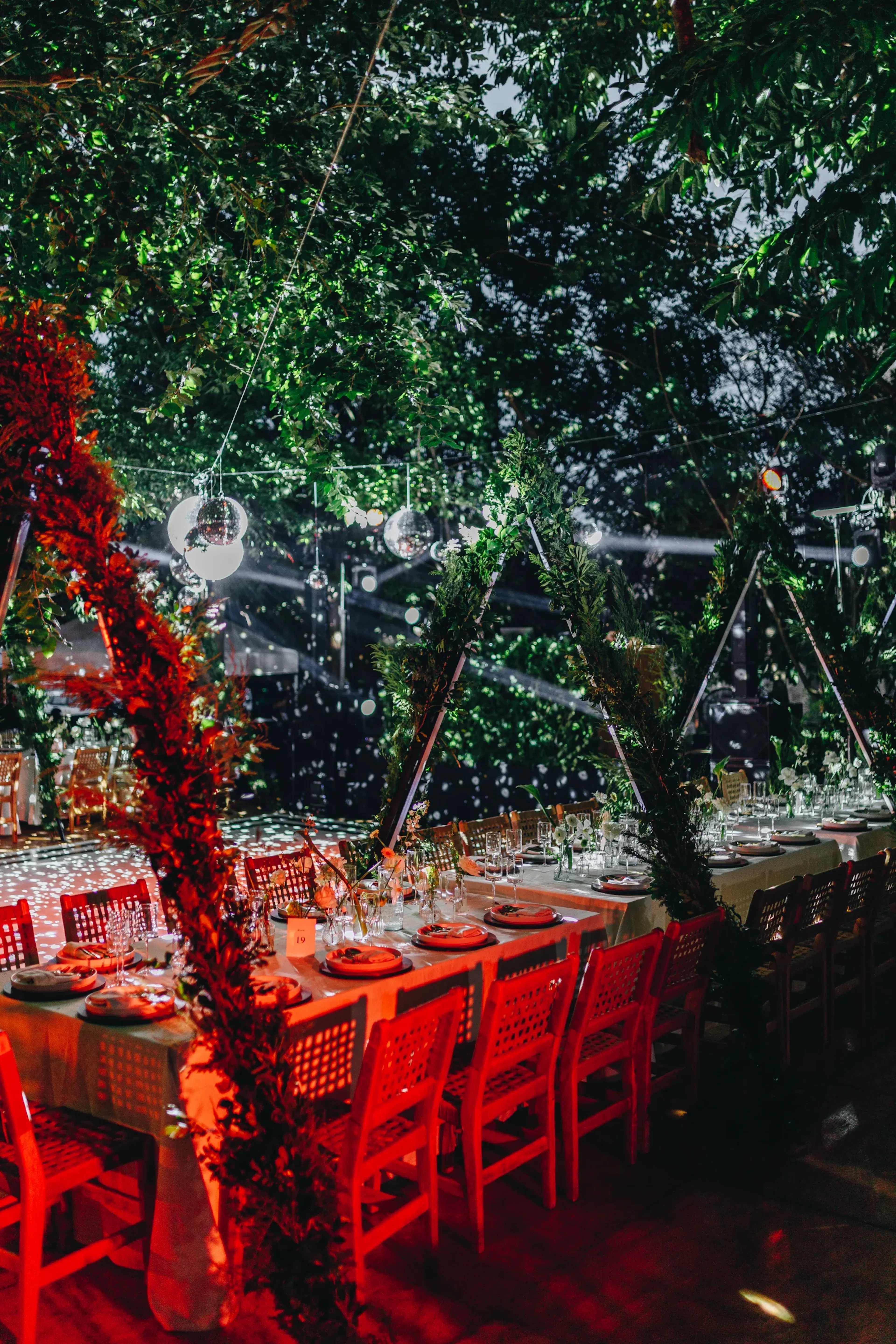 Cena festiva al aire libre con una mesa larga preparada para una fiesta. Sillas rojas y elegante cristalería bajo luces centelleantes y una exuberante decoración verde.