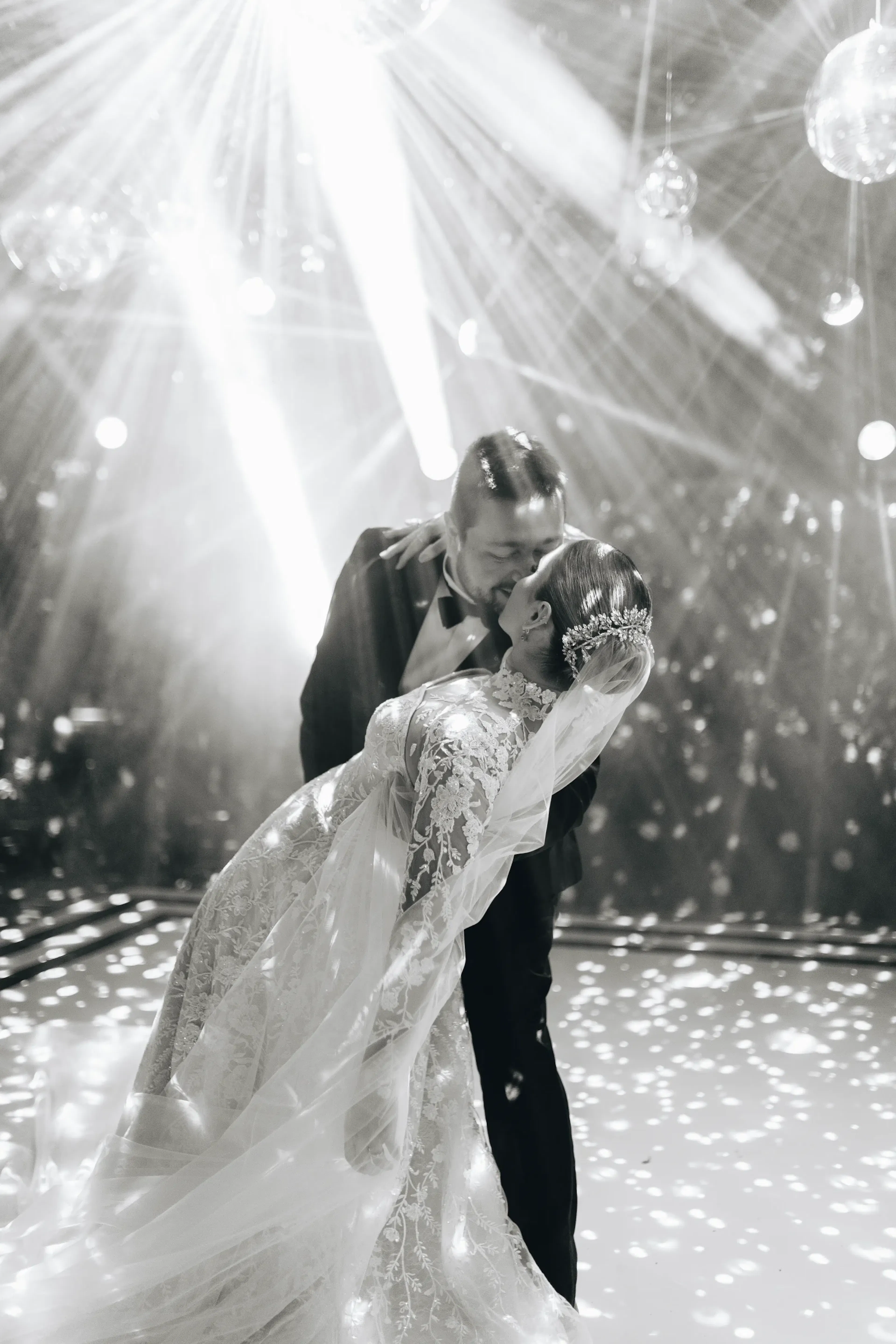 Una novia y un novio comparten un baile romántico bajo las brillantes luces de discoteca. Ella lleva un vestido ornamentado y están rodeados de rayos de luz que evocan alegría.