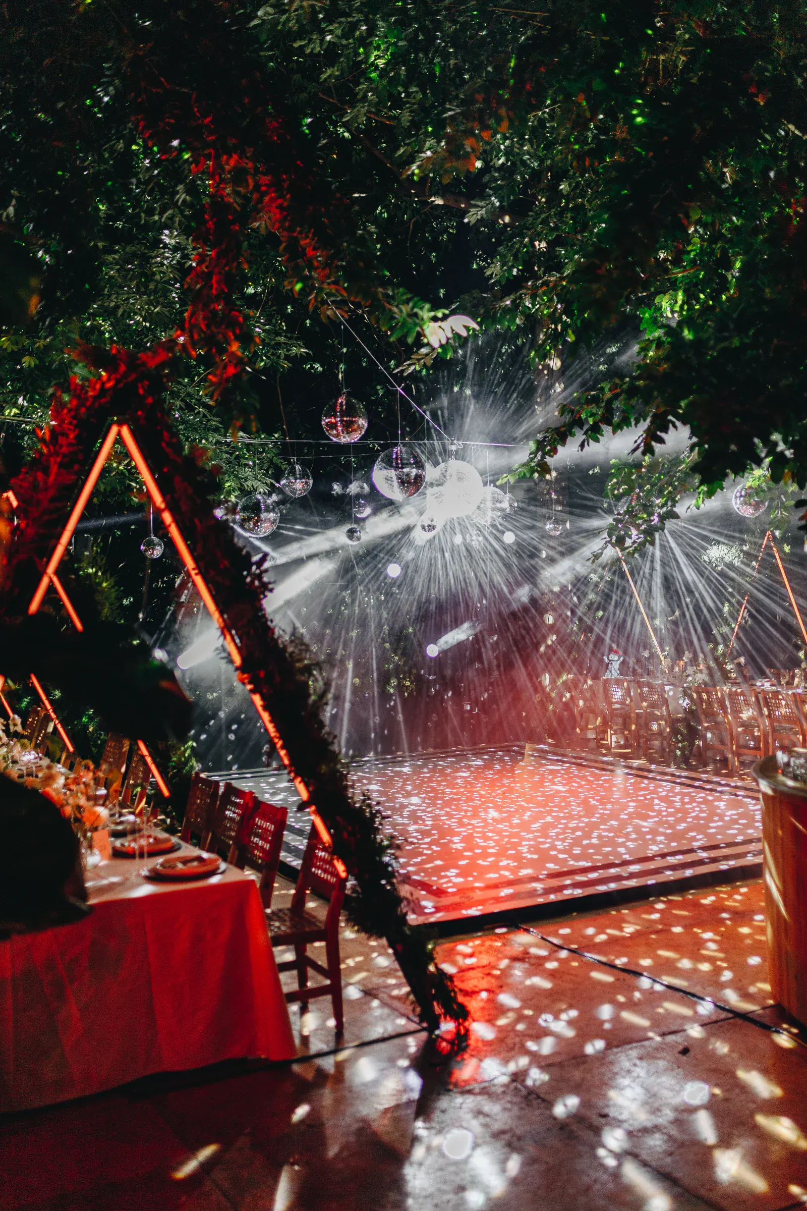 Una vibrante pista de baile iluminada con luces de discoteca rojas y blancas bajo un dosel de árboles en una fiesta al aire libre. Cerca hay mesas con manteles y sillas blancas.