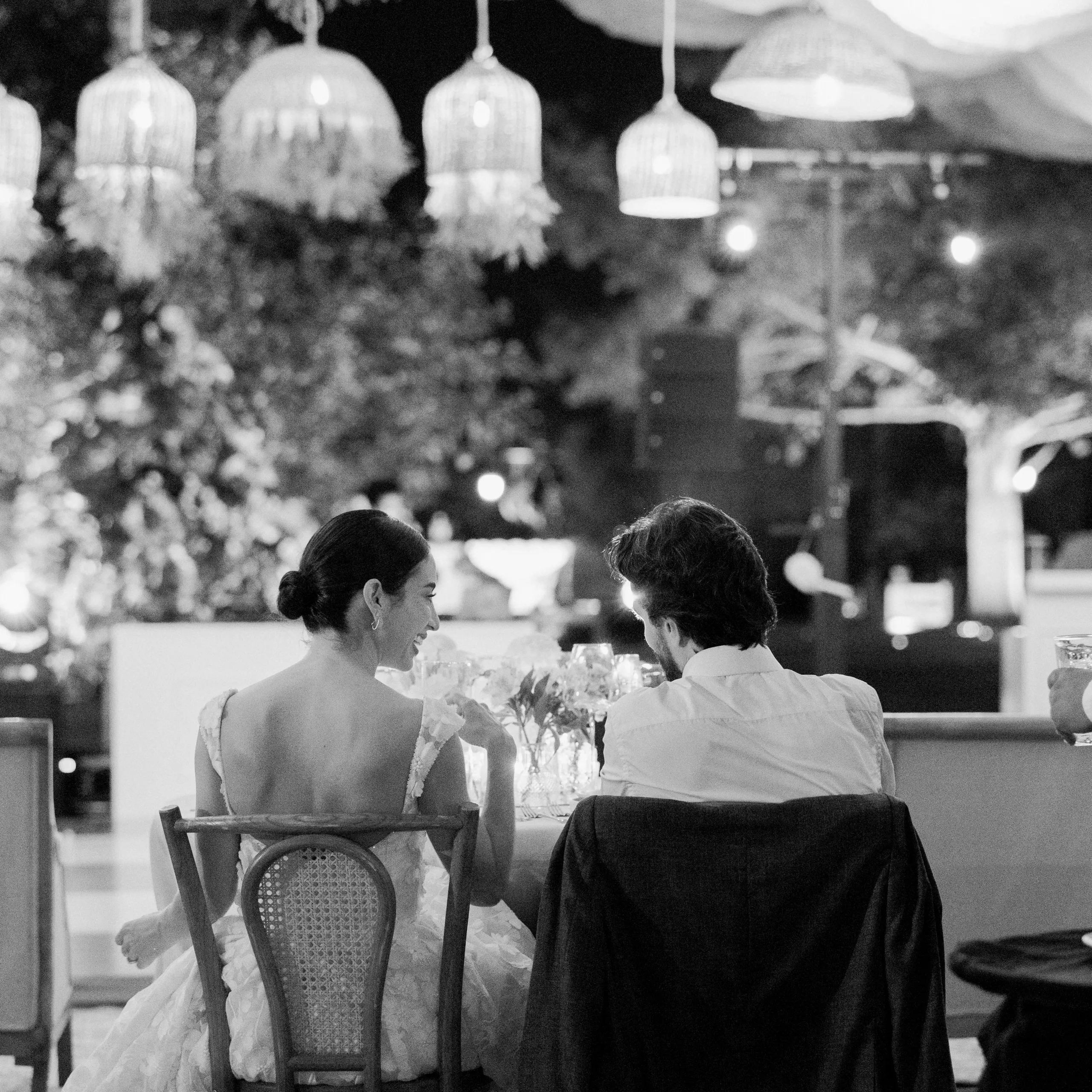 Una novia con un vestido de encaje y un novio con traje se sientan juntos en una mesa adornada con velas. La suave iluminación cenital y la decoración con hojas crean un ambiente romántico.