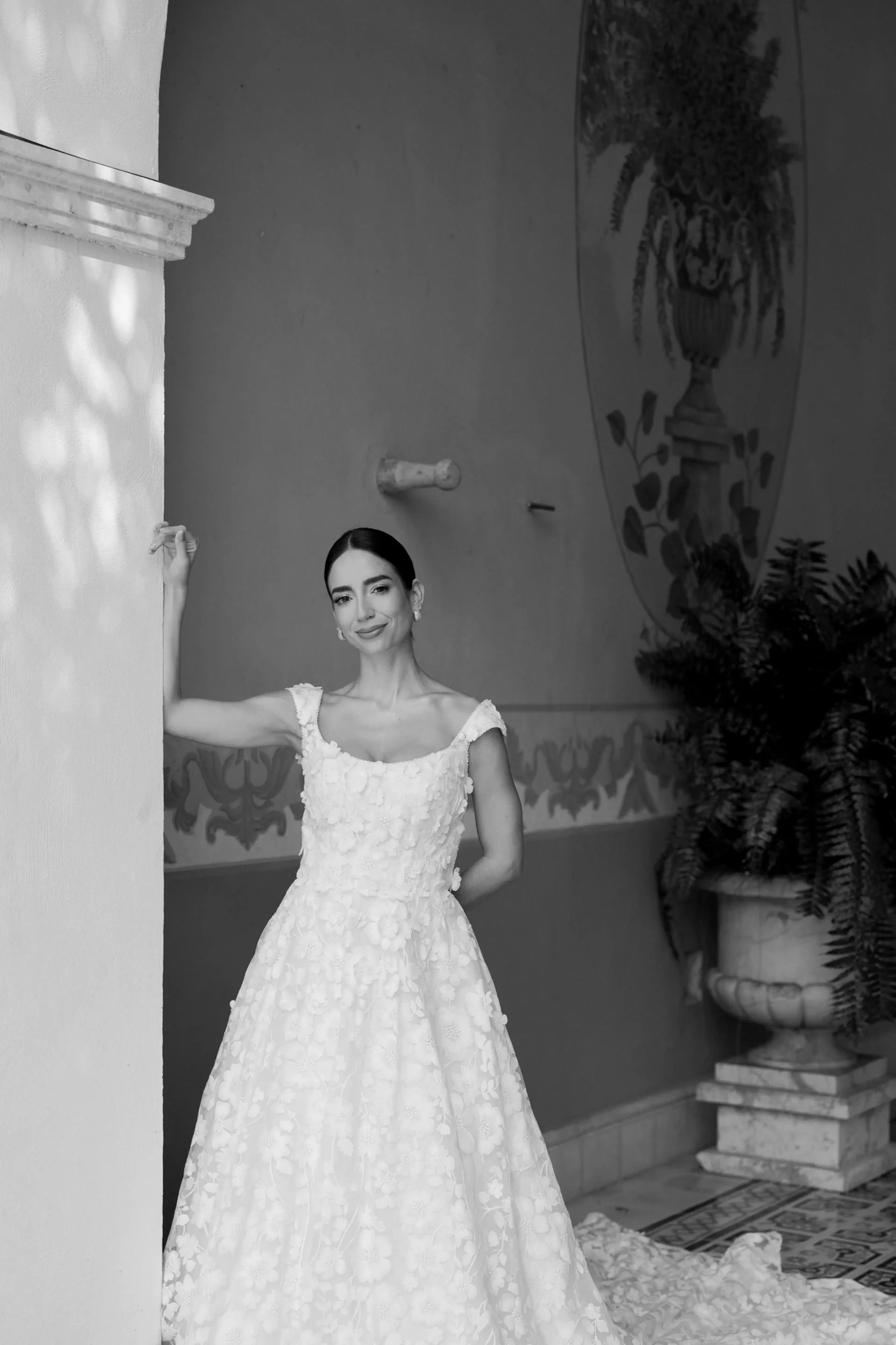 Una mujer con un elegante vestido blanco texturizado posa con gracia junto a una columna en un interior. El fondo presenta plantas decorativas, un mural y una iluminación tenue.