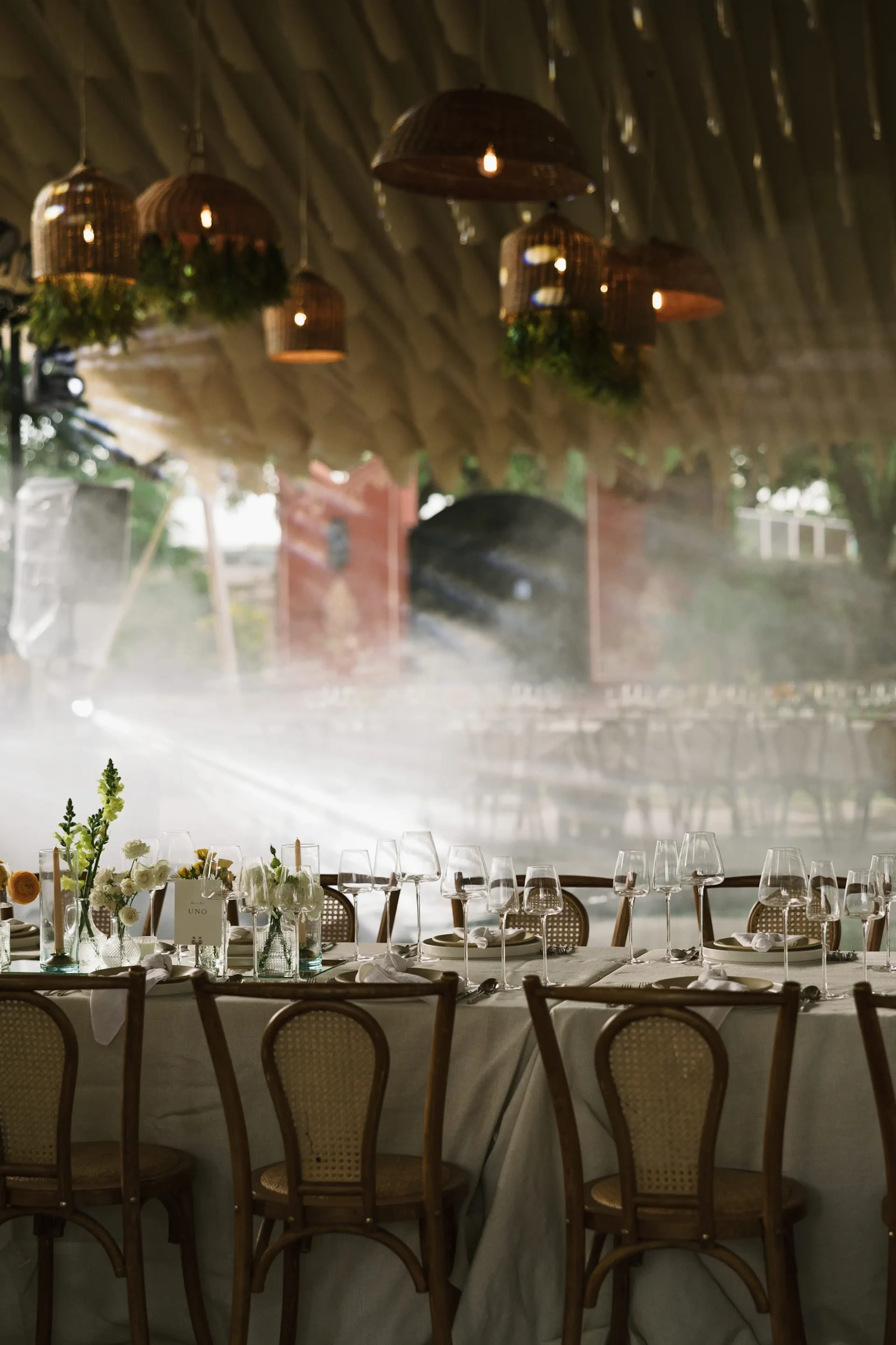 Elegante mesa con sillas de madera bajo lámparas colgantes de mimbre. Flores blancas, cubiertos y cristalería crean un ambiente diáfano y sofisticado.