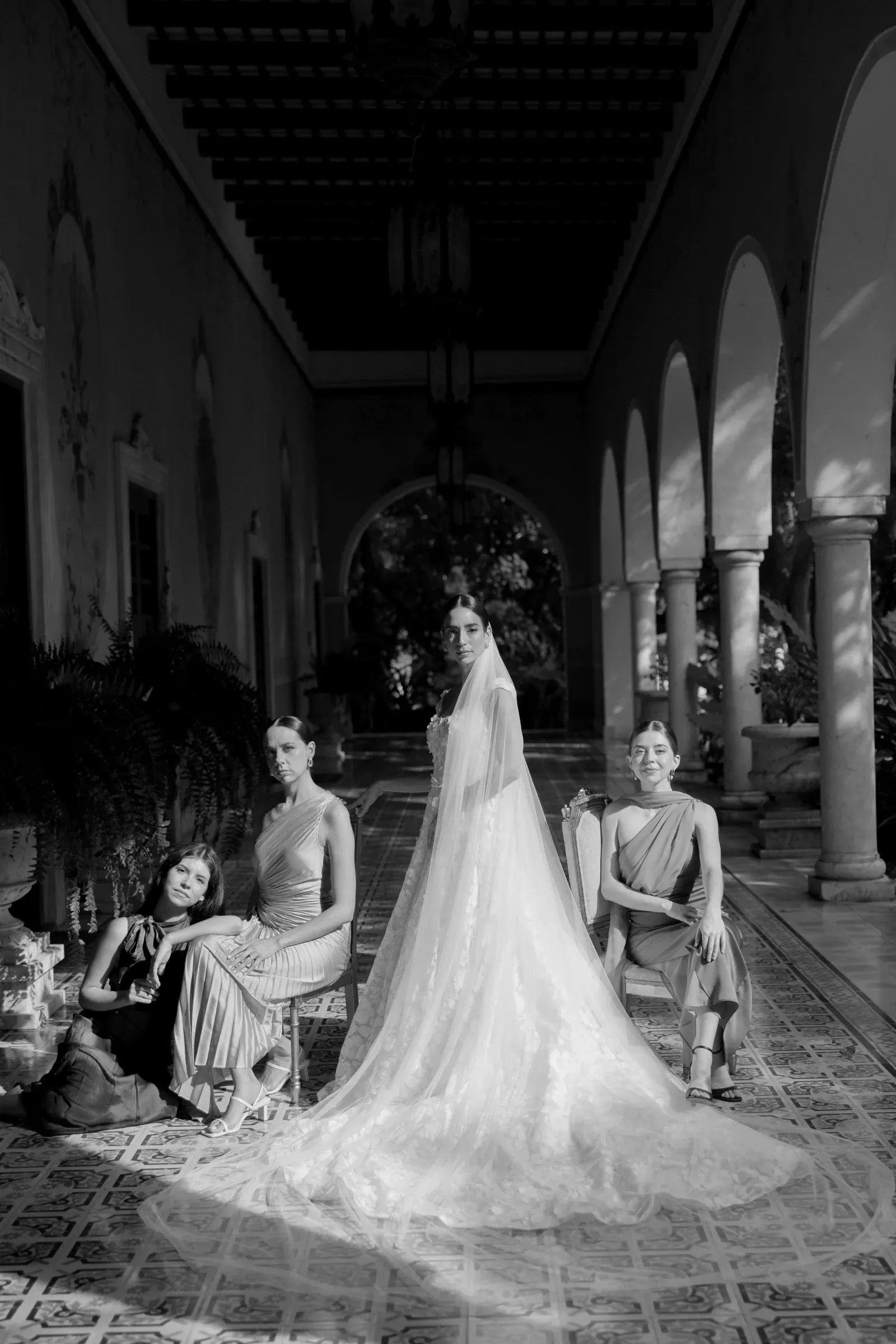 Elegante fotografía en blanco y negro de una novia con un velo suelto, de pie entre damas de honor sentadas en un pasillo arqueado y ornamentado, que transmite una gracia atemporal.