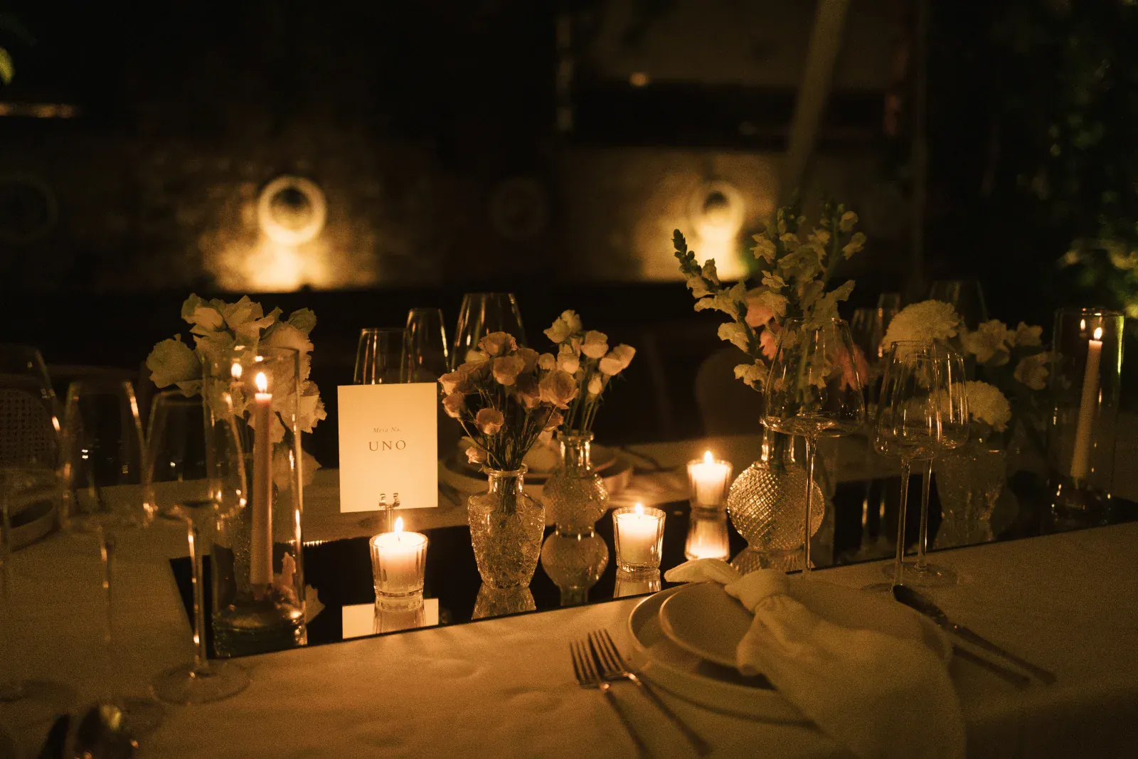 Elegante mesa de cena iluminada con velas para un evento, con jarrones con flores blancas y rosas, cristalería y una tarjeta de menú, creando un ambiente cálido e íntimo.