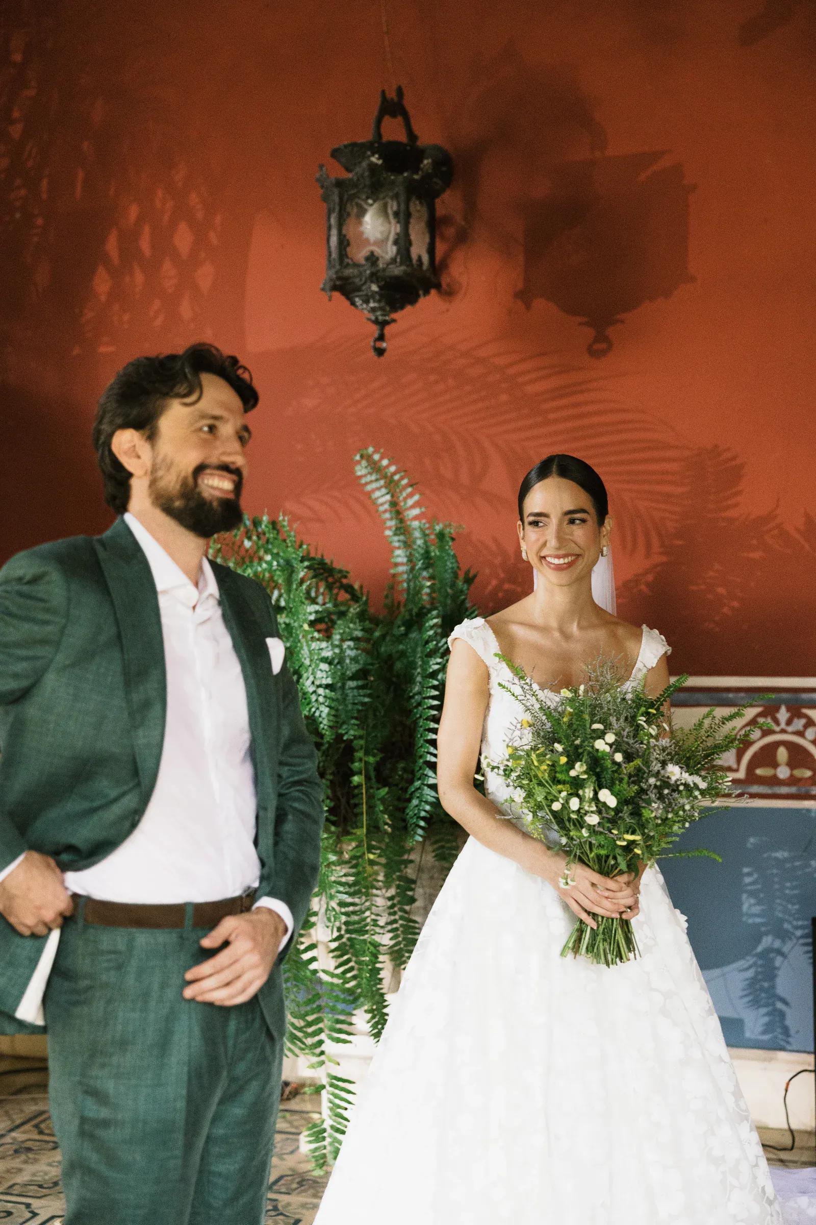 Una novia con un vestido blanco sostiene un ramo de flores silvestres y sonríe cálidamente a un hombre con traje verde. Se encuentran frente a una pared roja con plantas decorativas, transmitiendo alegría.