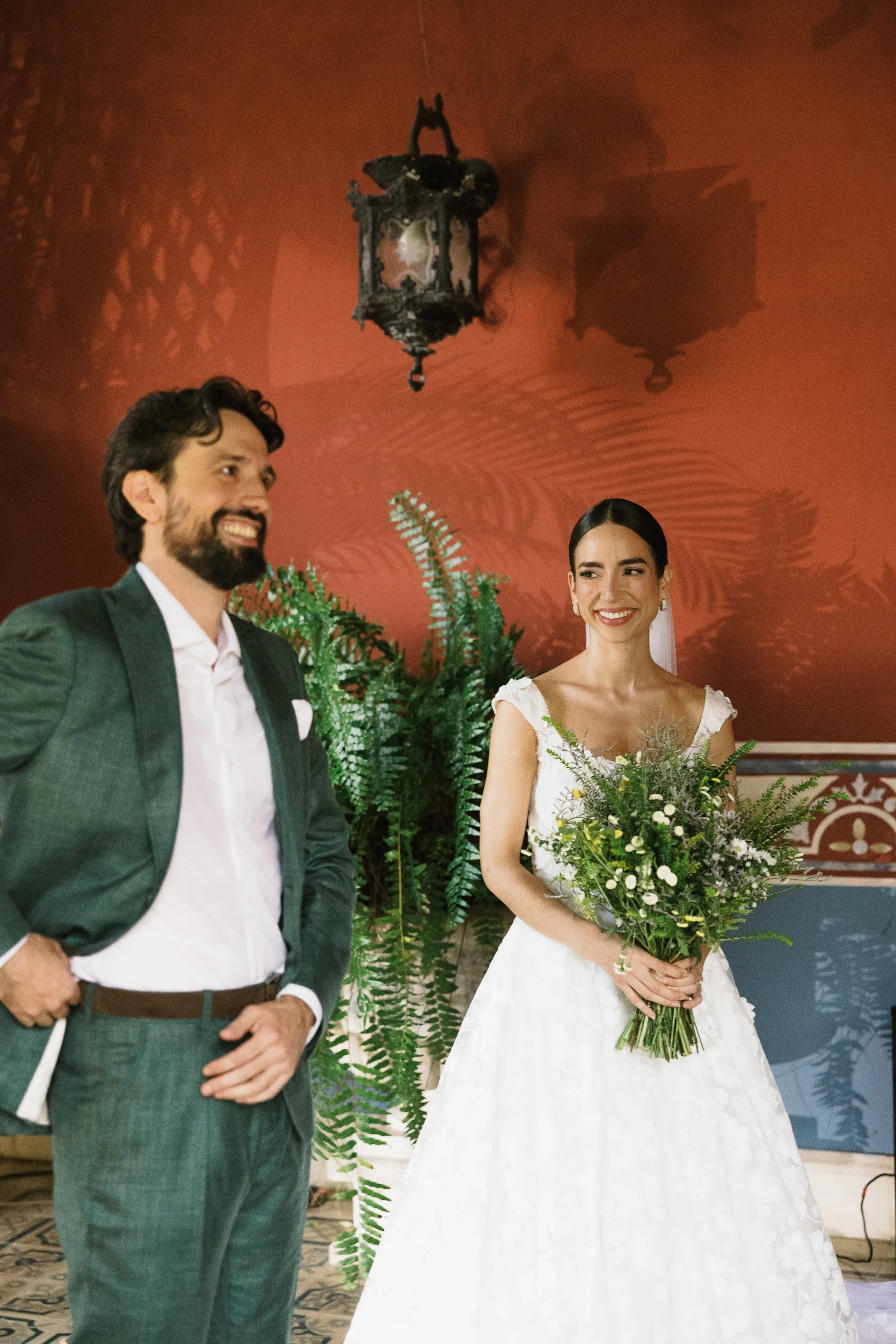Una novia con un vestido blanco sostiene un ramo de flores silvestres y sonríe cálidamente a un hombre con traje verde. Se encuentran frente a una pared roja con plantas decorativas, transmitiendo alegría.