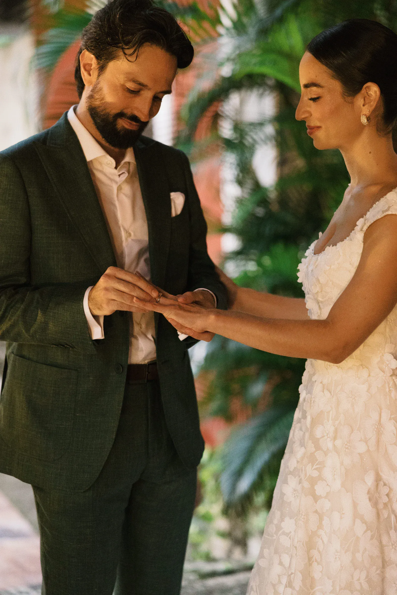 Un novio con traje verde le pone un anillo a la novia. Ella lleva un vestido blanco de flores. Se encuentran en un jardín, transmitiendo alegría y amor.