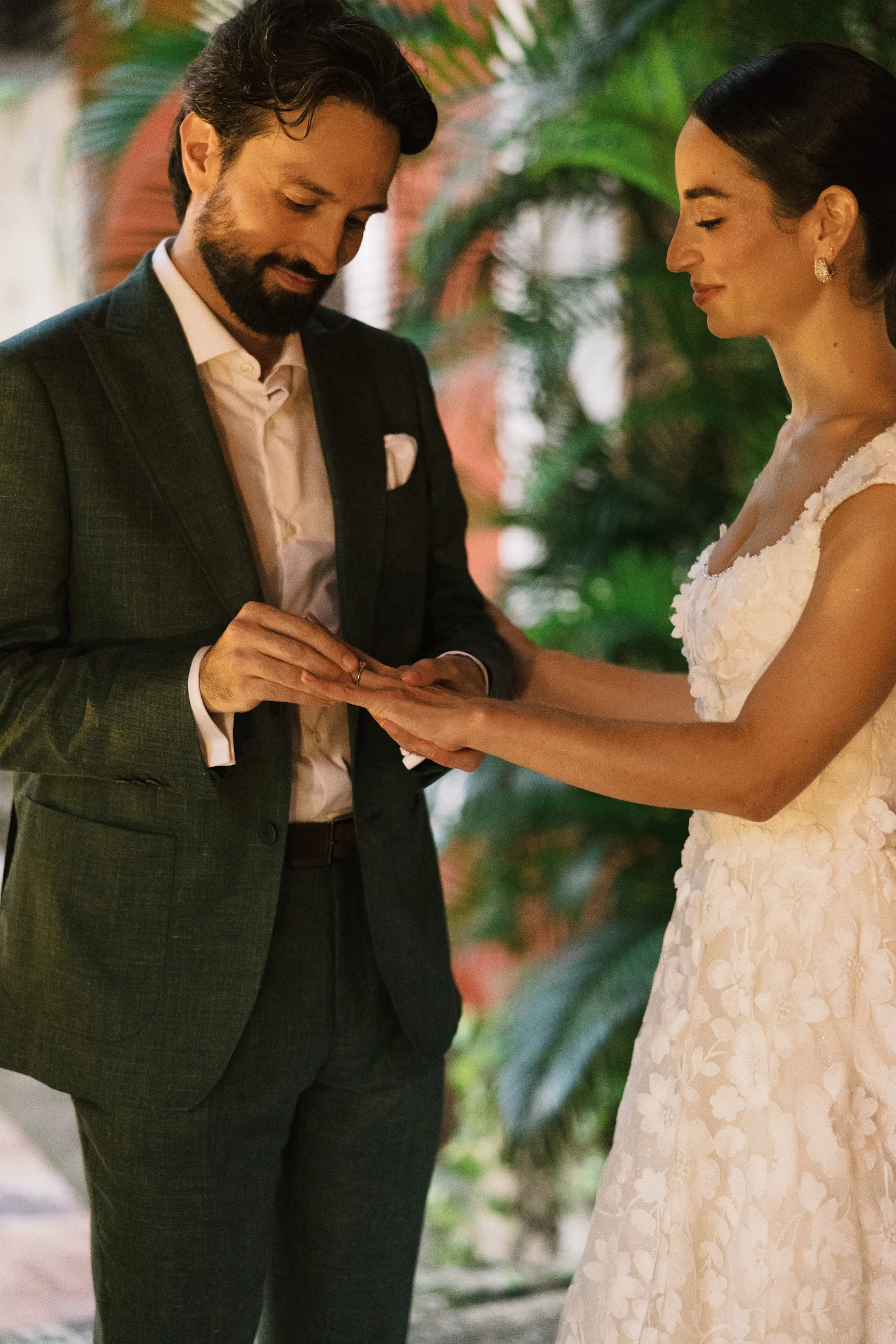 Un novio con traje verde le pone un anillo a la novia. Ella lleva un vestido blanco de flores. Se encuentran en un jardín, transmitiendo alegría y amor.
