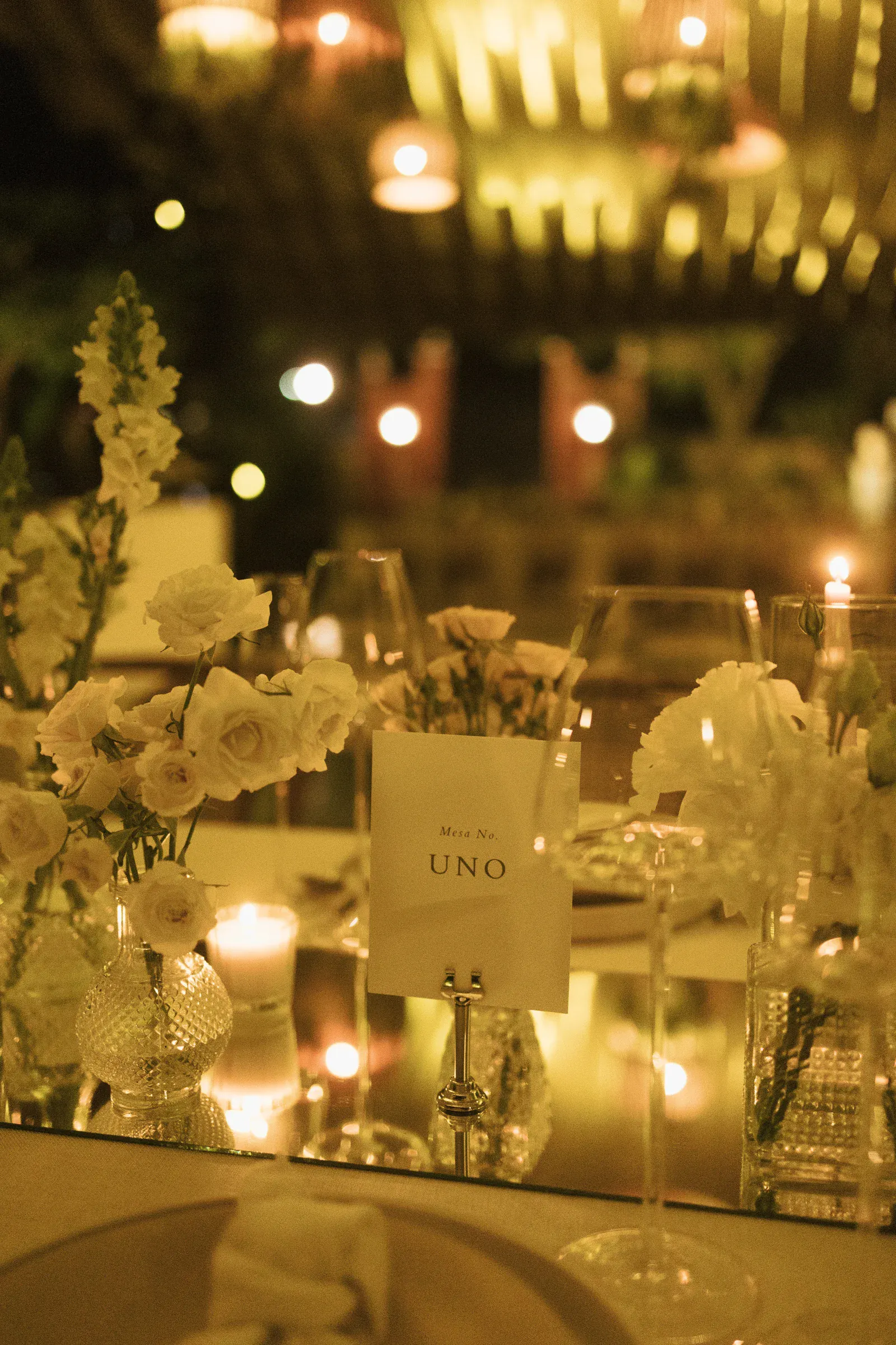 Elegante mesa dispuesta en un evento con poca luz, con flores blancas en jarrones, velas, copas de vino y una tarjeta con la leyenda "Mesa No. UNO", creando un ambiente acogedor.