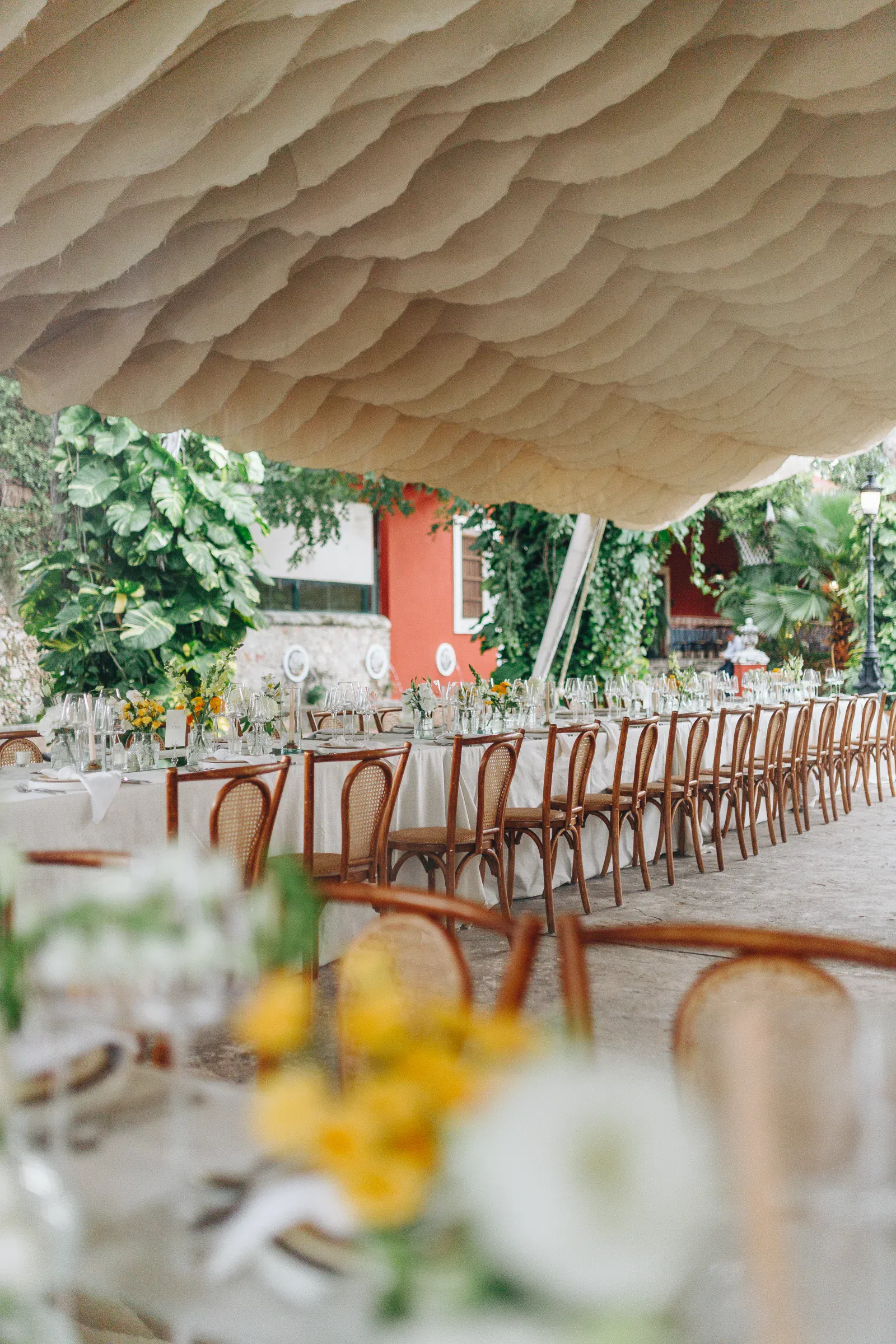 Una larga mesa de comedor al aire libre con sillas de madera, preparada para un evento bajo un dosel ondulado. Rodeada de exuberante vegetación y un edificio rojo, crea un ambiente festivo.