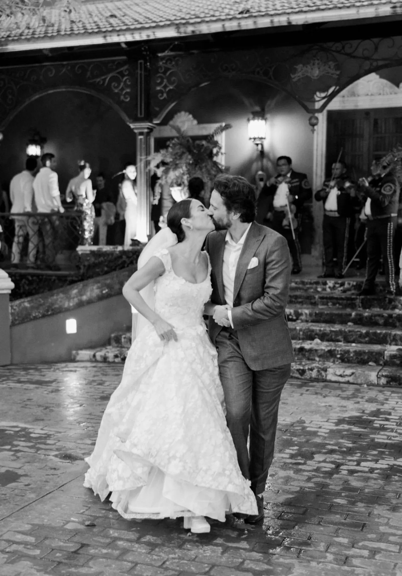 Una novia y un novio se besan alegremente a la salida de un local. La novia lleva un vestido de encaje y el novio, un traje. Al fondo, se ven invitados y músicos.