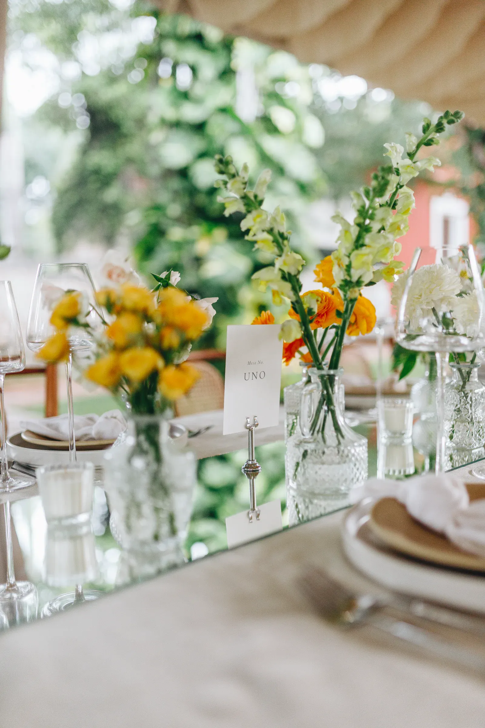 Elegante mesa con superficie espejada, con flores amarillas y naranjas en jarrones de vidrio, platos cuidadosamente dispuestos y copas de vino, creando una atmósfera fresca y acogedora.
