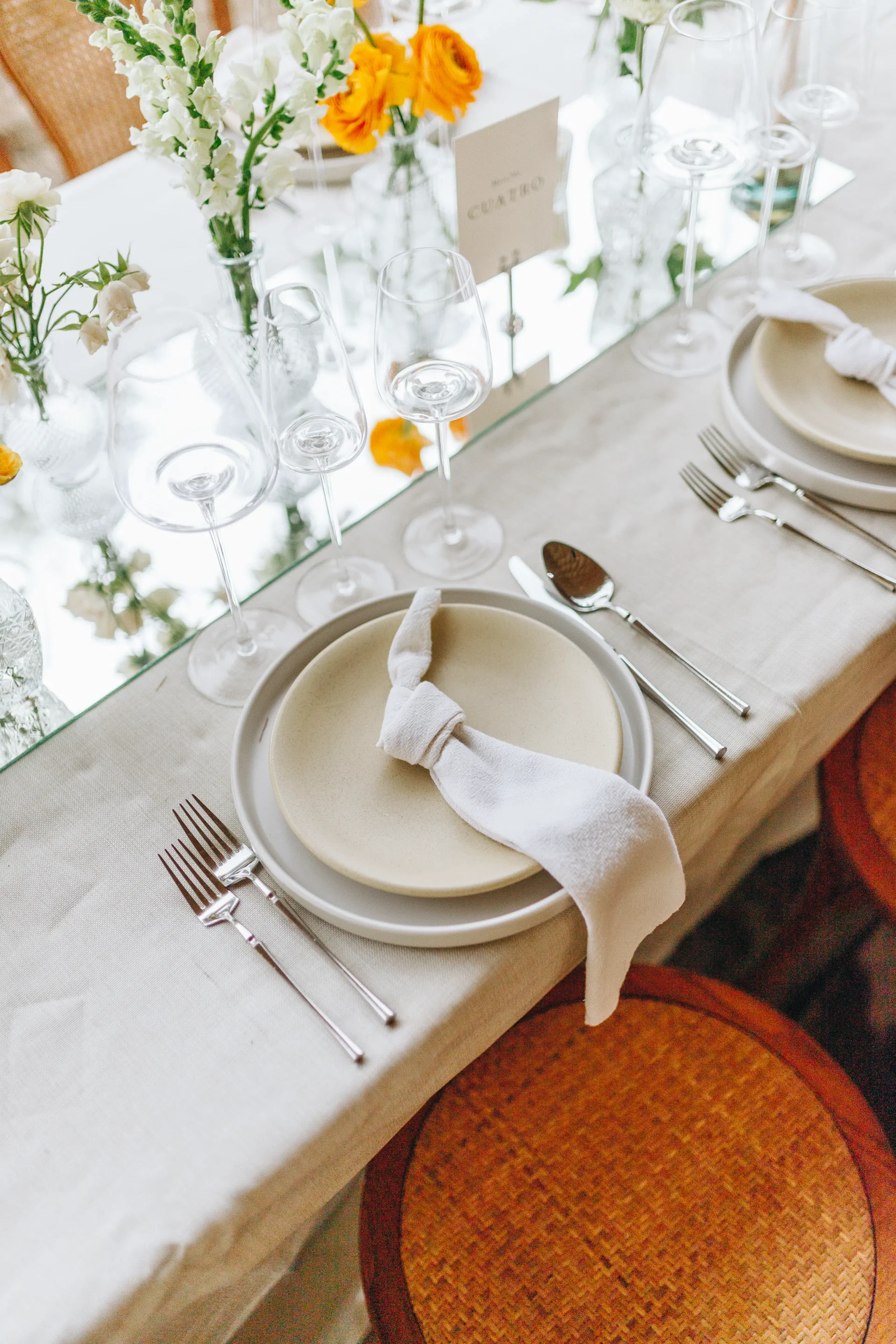 Elegante mesa con platos blancos y servilletas de tela anudadas, rodeada de copas de vino. Flores de color naranja brillante y blanco aportan un toque fresco.