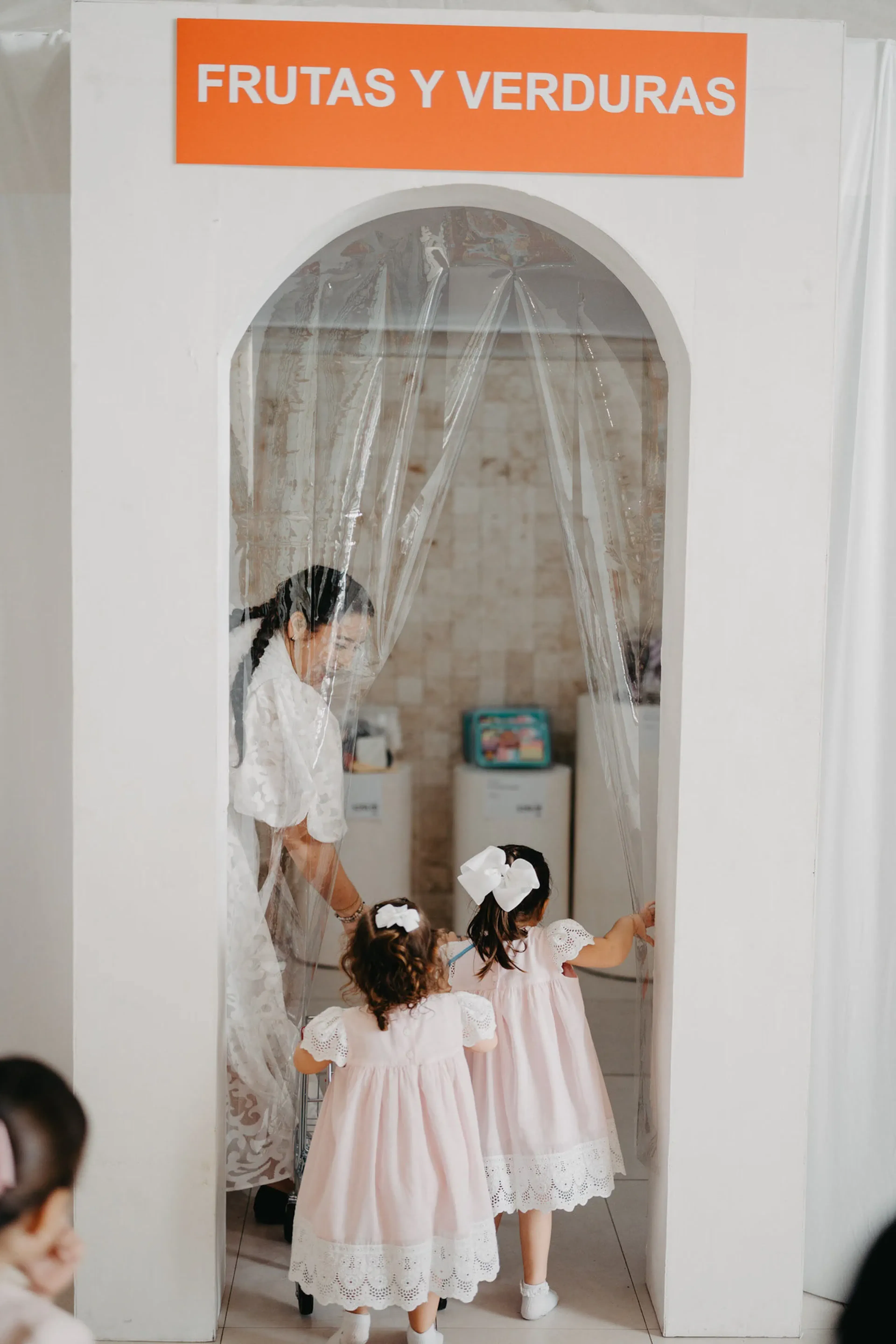 Una mujer guía a dos niñas a través de un arco blanco con la inscripción "Frutas y Verduras". Las niñas, con vestidos rosas iguales, irradian curiosidad y alegría.