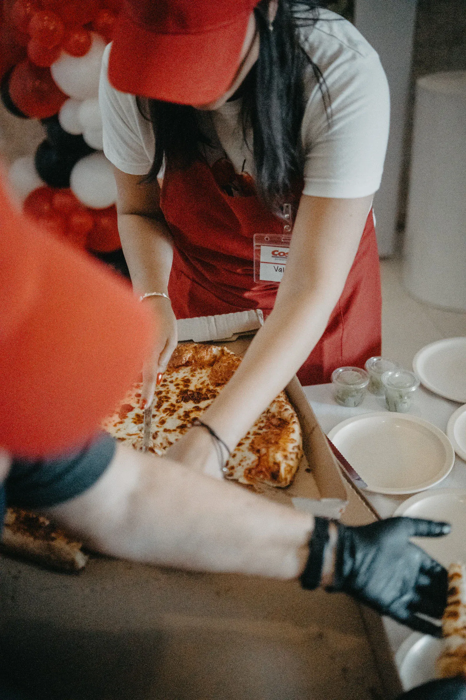 Una persona con delantal y gorra rojos corta una pizza grande de queso en una caja de cartón. Cerca, se colocan platos y vasitos, con globos rojos y blancos de fondo.