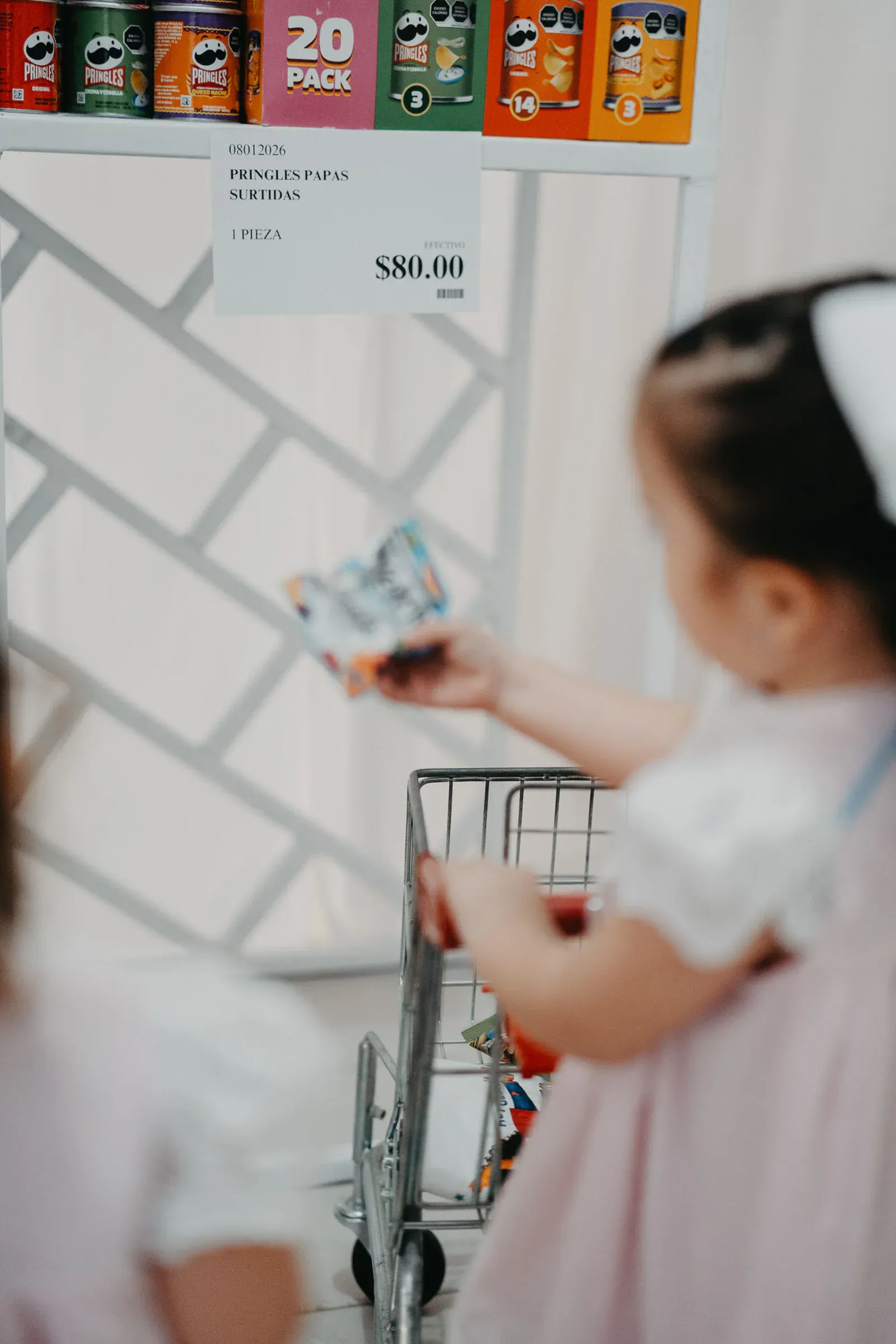 Una niña pequeña con vestido rosa y lazo blanco compra con un carrito de juguetes, buscando un paquete de refrigerios. El estante exhibe coloridas latas de Pringles con una etiqueta de precio.