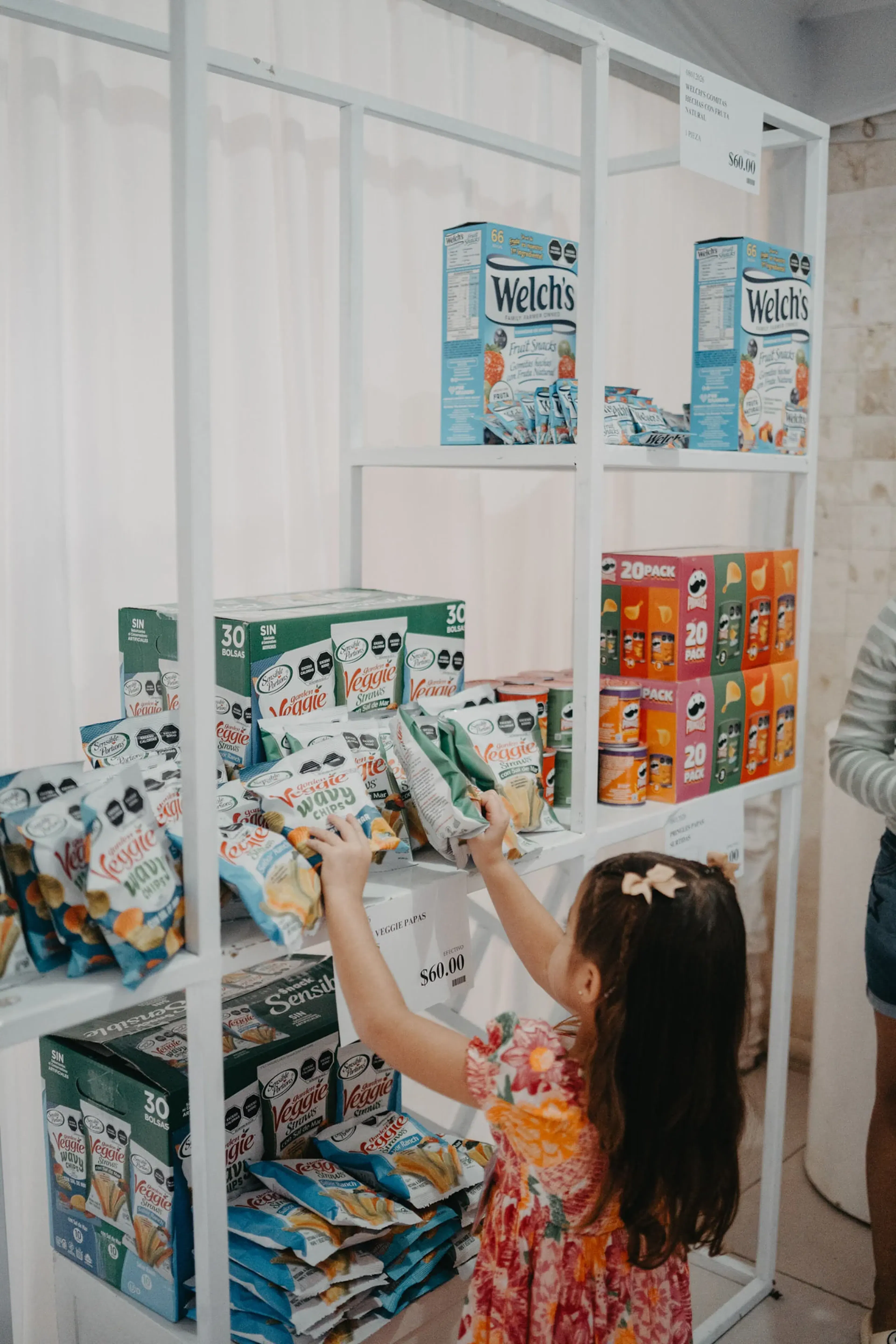 Una joven con un vestido floreado busca paquetes de bocadillos en un estante lleno de diversos alimentos envasados, lo que transmite curiosidad e interés.
