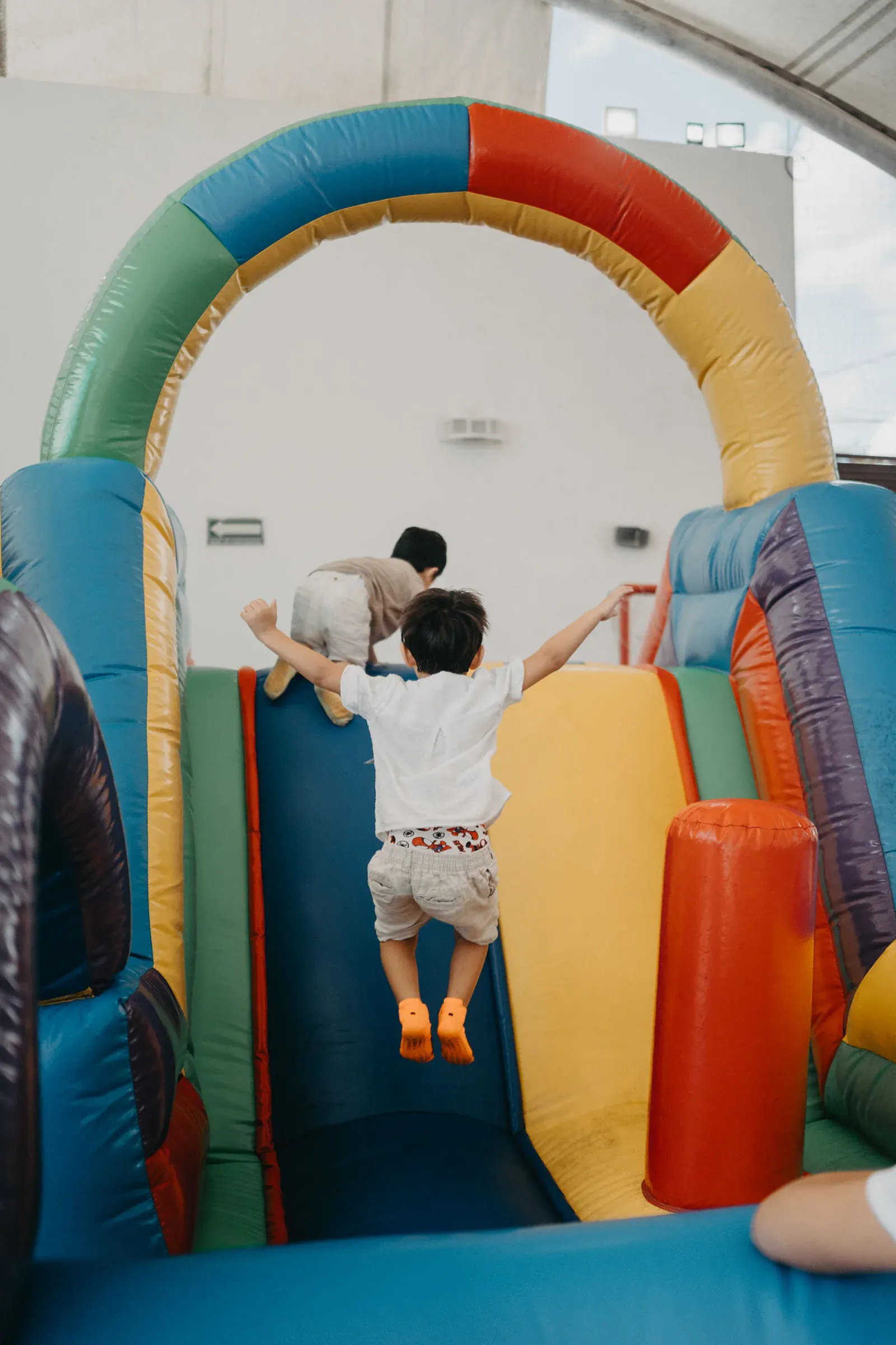 Un niño salta alegremente en un tobogán inflable de colores, con los brazos extendidos. Otro niño trepa delante. El ambiente es lúdico y animado.