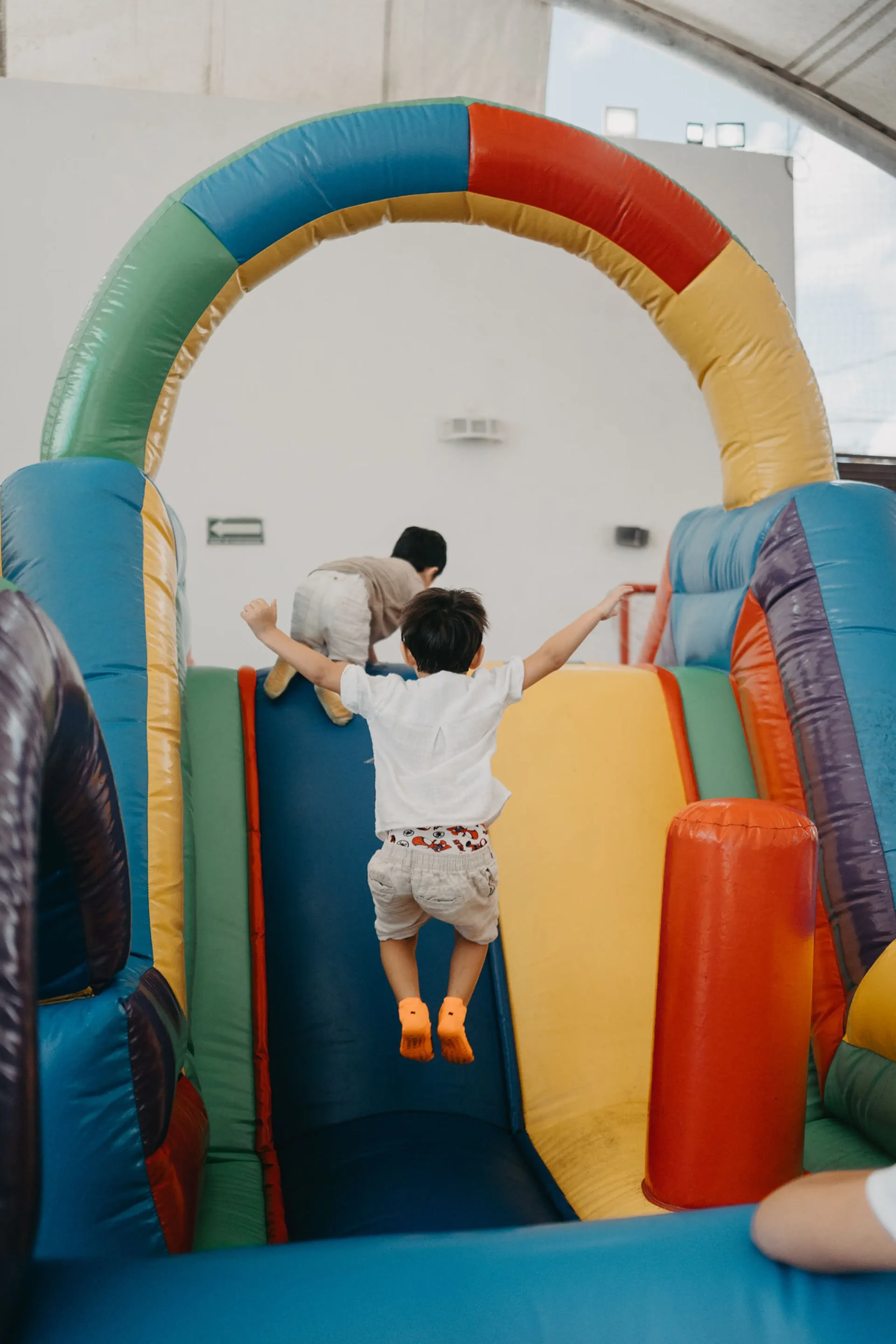 Un niño salta alegremente en un tobogán inflable de colores, con los brazos extendidos. Otro niño trepa delante. El ambiente es lúdico y animado.