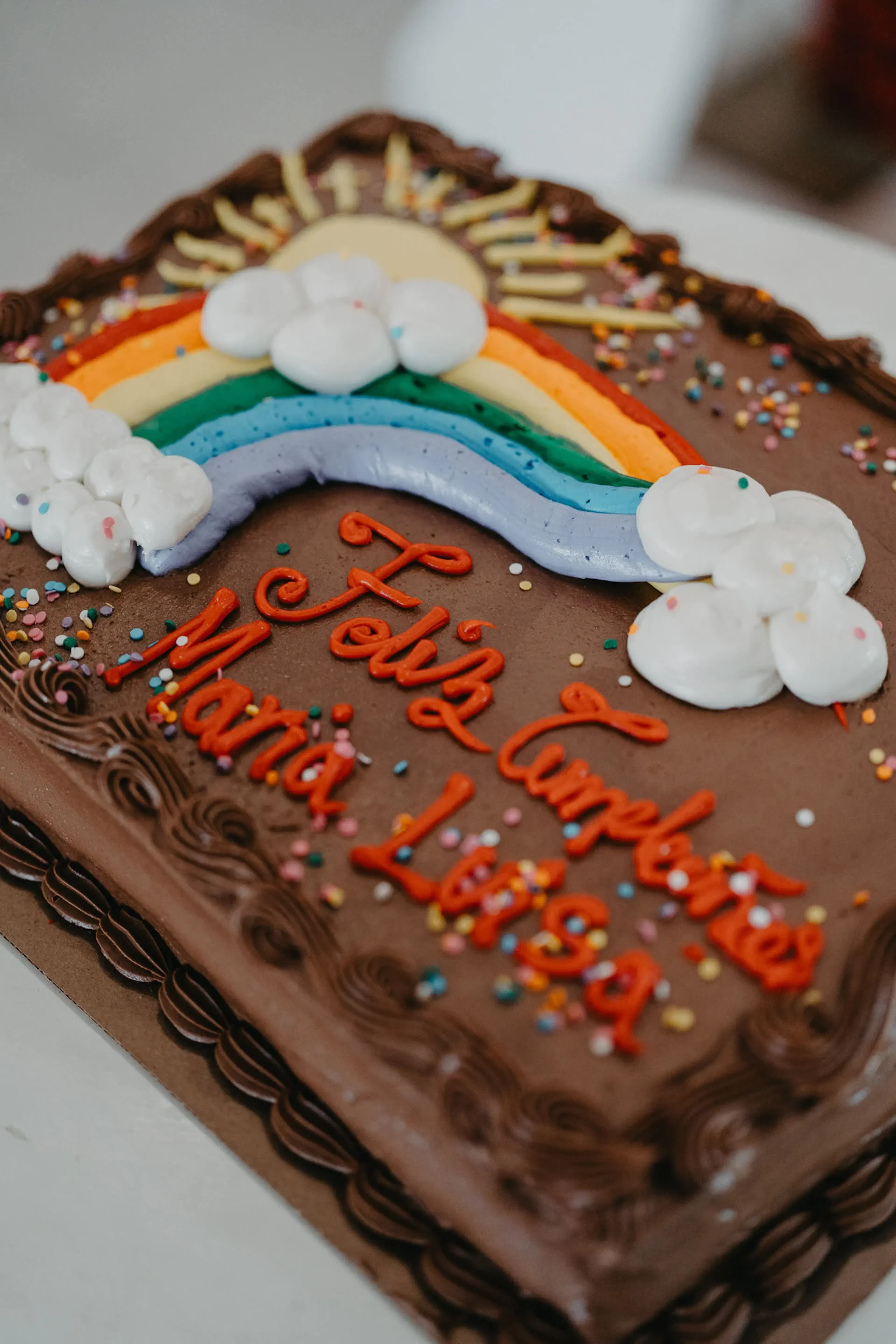 Pastel de chocolate con un colorido diseño de arcoíris y suaves nubes blancas. "Feliz Cumpleaños María Luisa" está escrito con glaseado rojo, rodeado de chispitas.