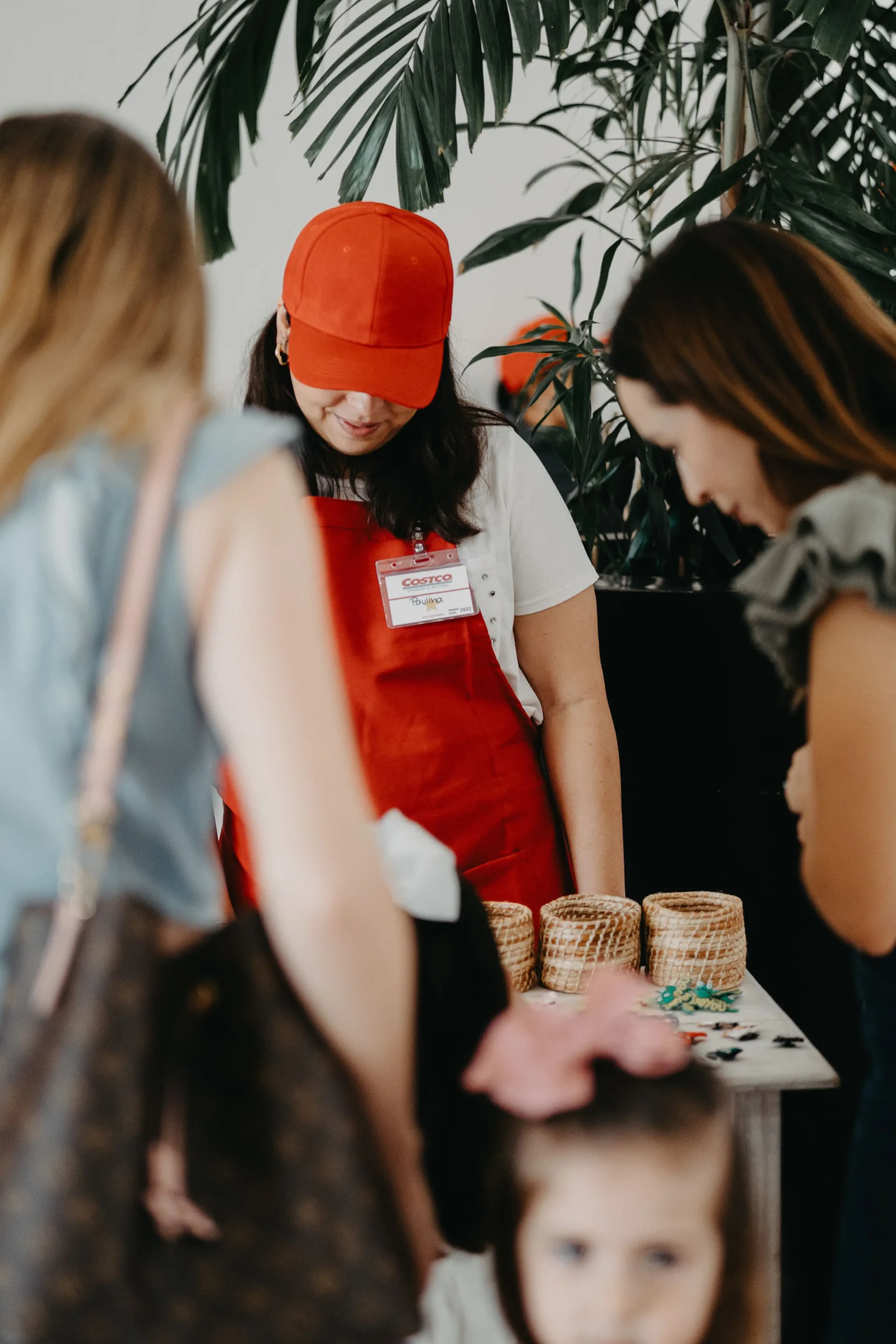 Una mujer con delantal y cofia rojos está de pie detrás de una mesa con cestas tejidas, conversando con los invitados. Está rodeada de vegetación, creando un ambiente cálido.