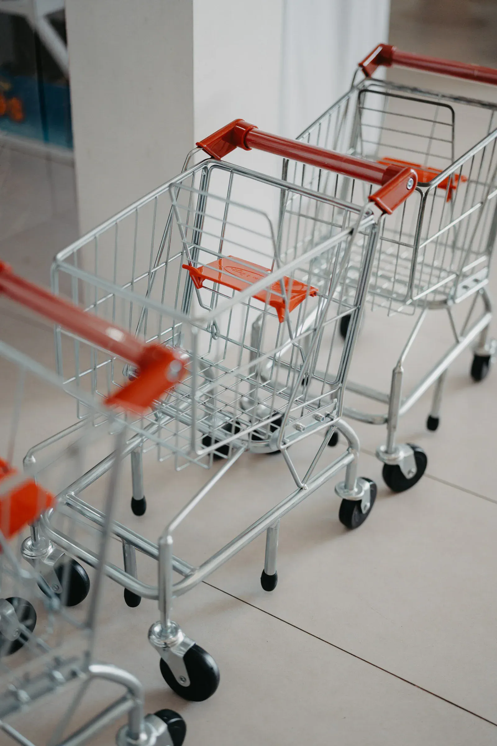 Tres pequeños carritos de compra metálicos con asas rojas se encuentran alineados sobre un suelo de baldosas, transmitiendo una sensación de organización y preparación.