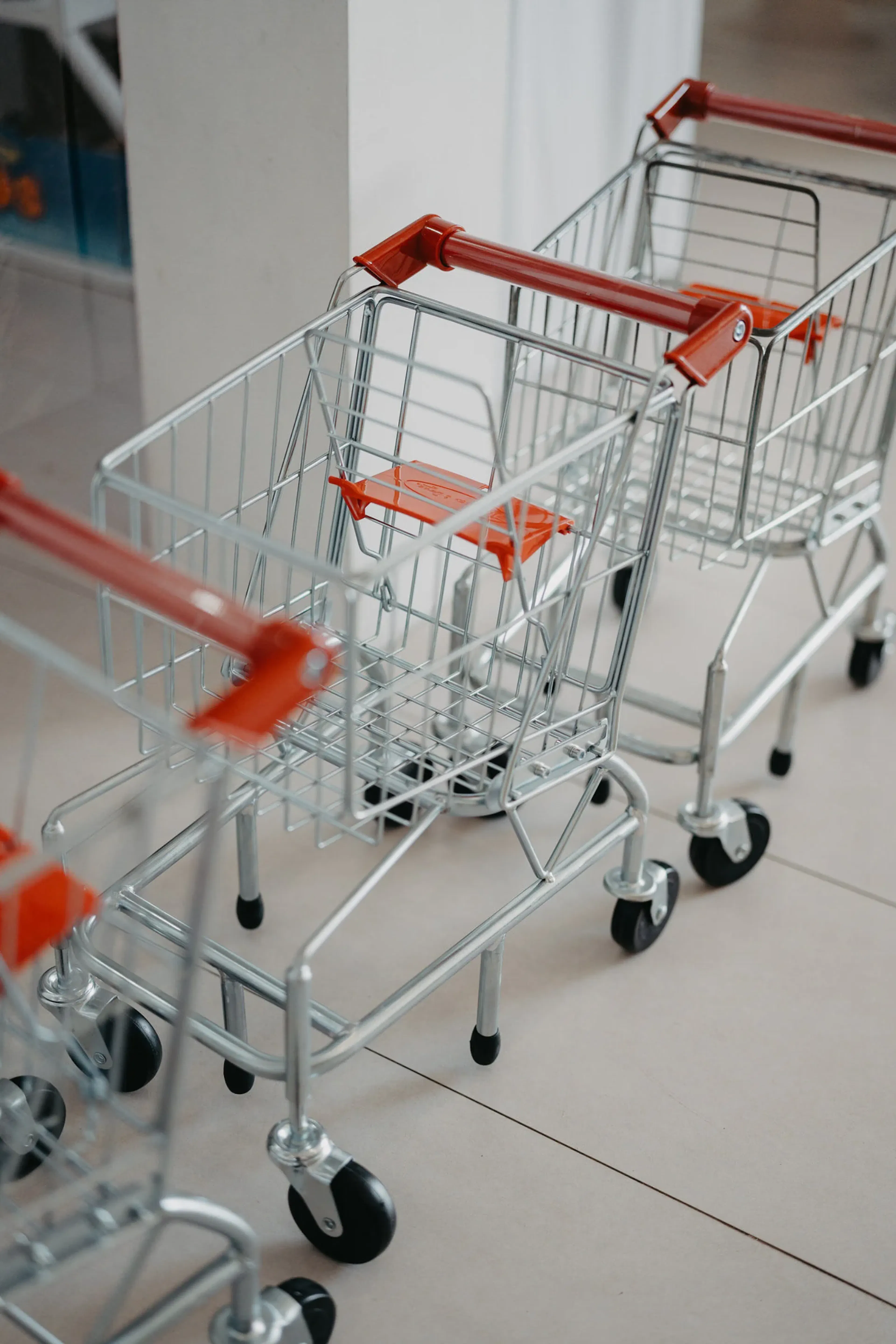 Tres pequeños carritos de compra metálicos con asas rojas se encuentran alineados sobre un suelo de baldosas, transmitiendo una sensación de organización y preparación.