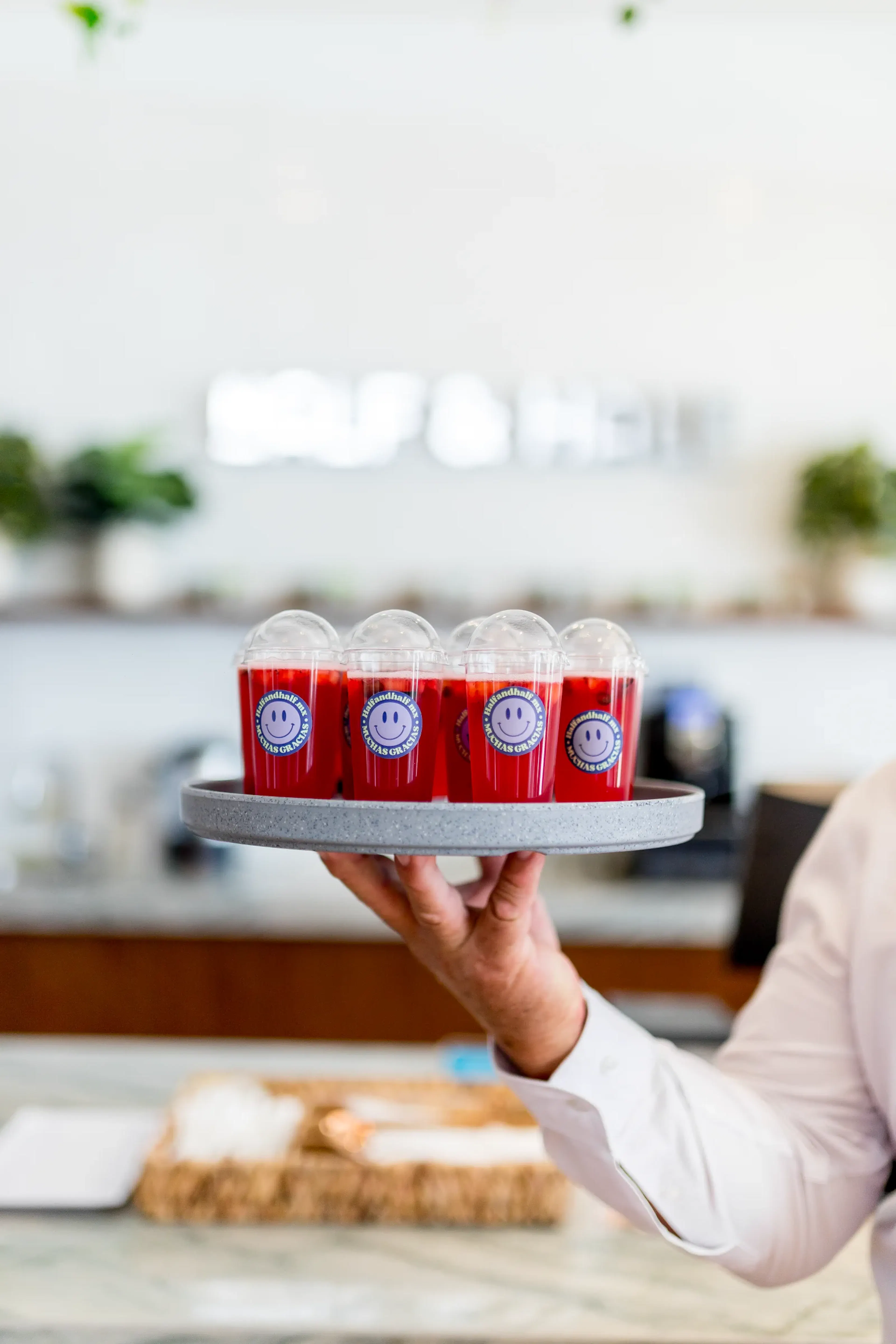 Una mano sostiene una bandeja con cinco bebidas rojas en vasos de plástico, cada una con una pegatina de una cara sonriente. El fondo es un café brillante y desenfocado.