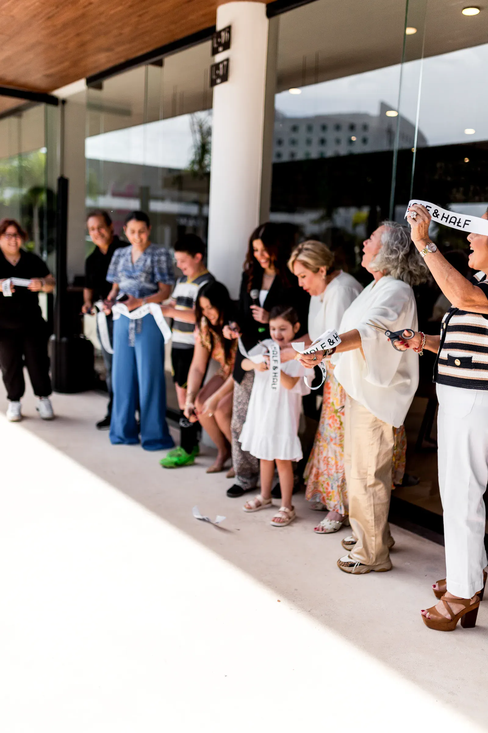 Un grupo diverso de personas, incluido un niño, participa en una ceremonia de inauguración afuera de un edificio moderno, lo que transmite una sensación de celebración y comunidad.