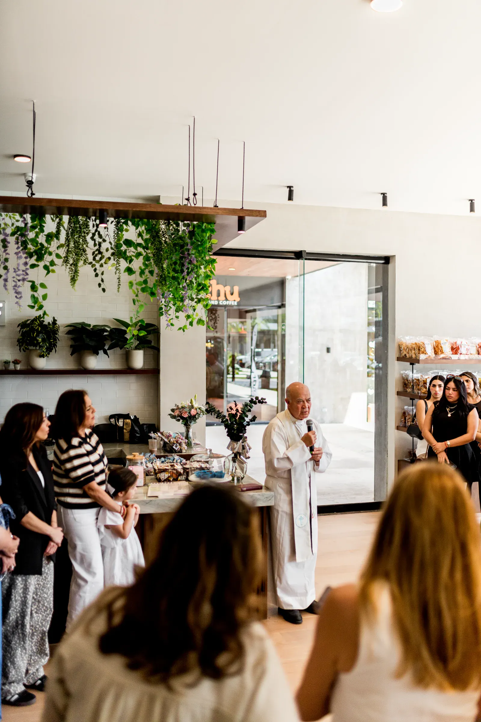 Un sacerdote habla en un café moderno con plantas verdes y estanterías colgantes. Un pequeño grupo escucha atentamente, creando un ambiente cálido e íntimo.