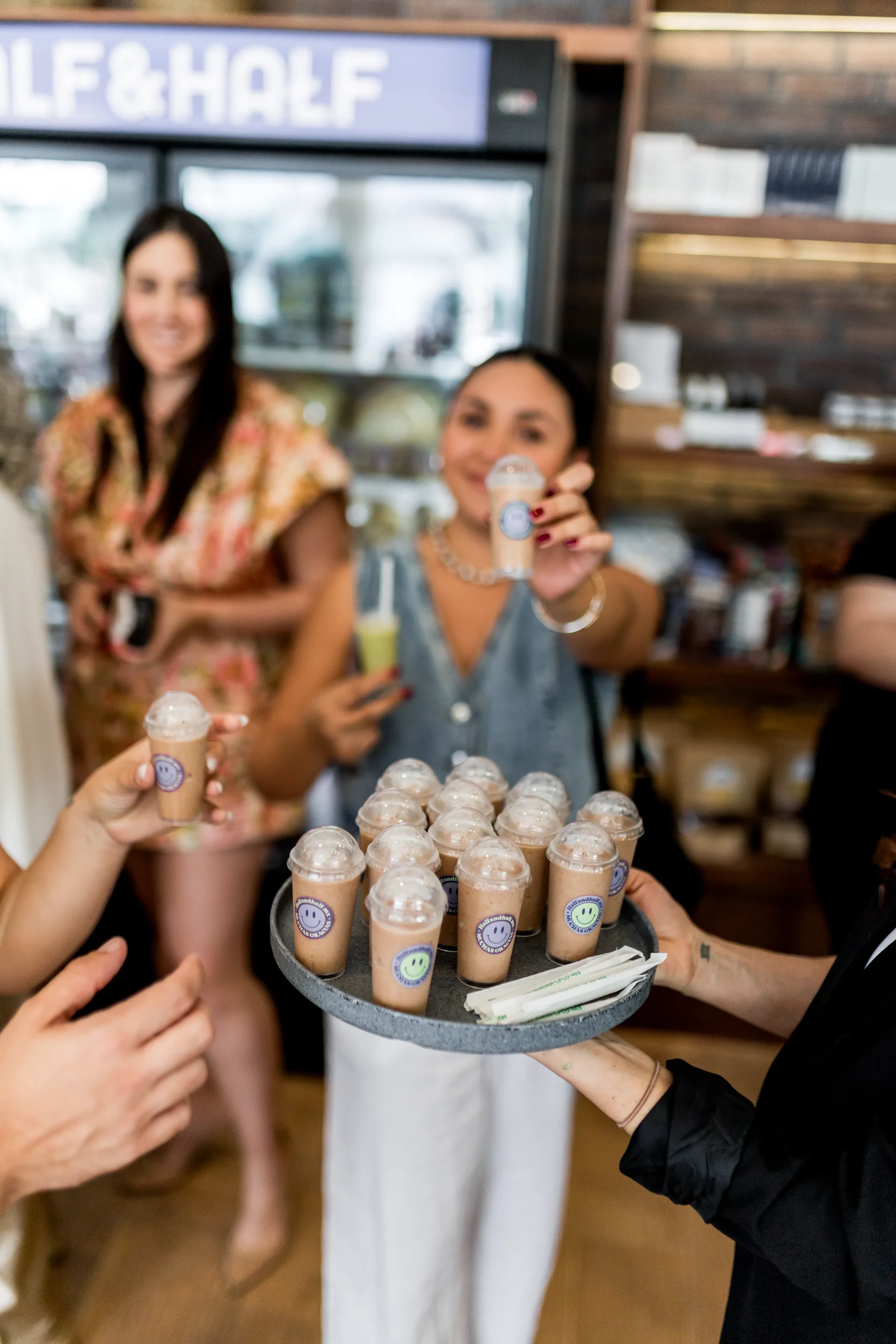 Un grupo de personas en una cafetería sostiene alegremente pequeñas y espumosas bebidas de café helado en una bandeja. El ambiente es animado, con un ambiente cálido y acogedor.
