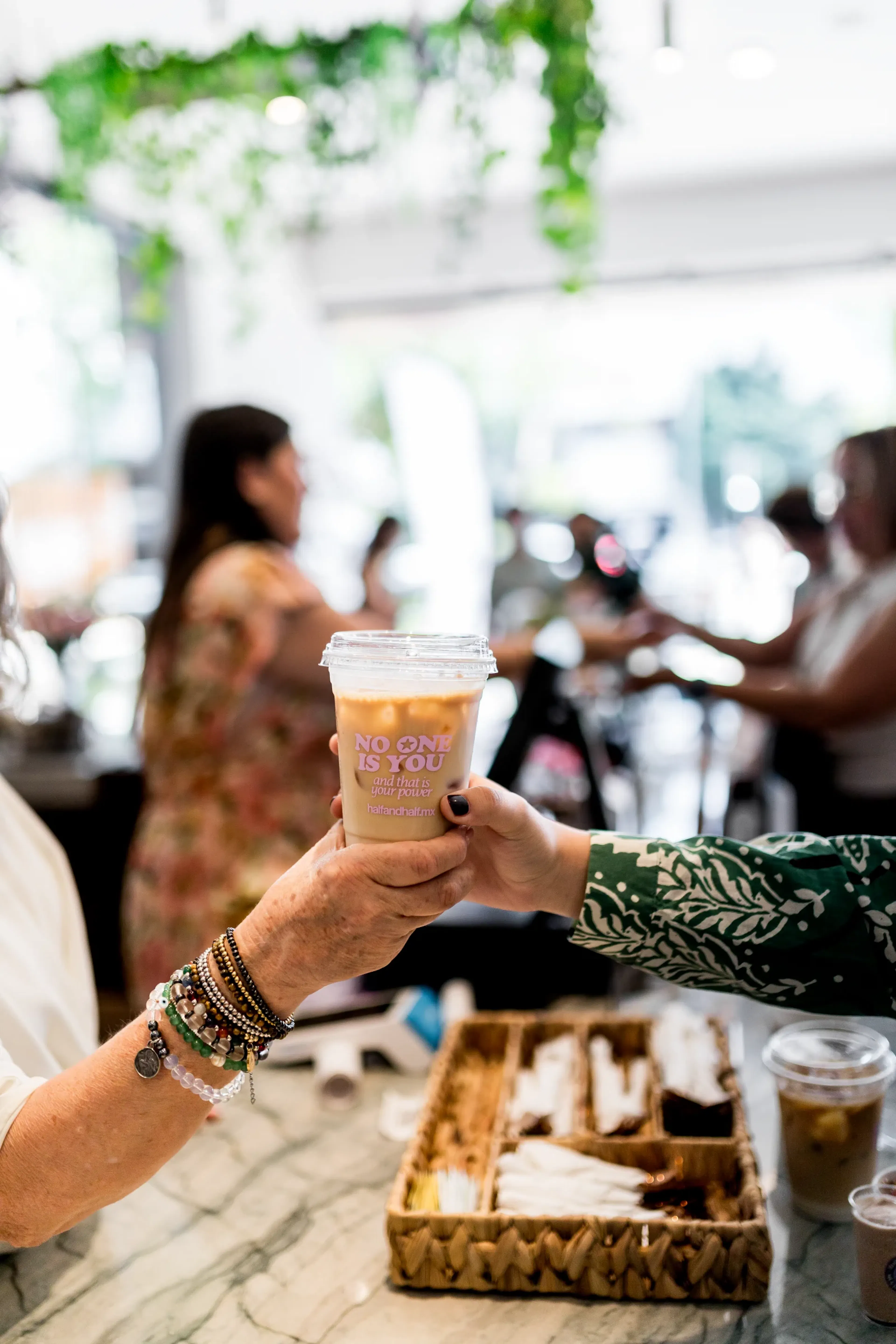 Dos personas intercambian una bebida en un café rodeado de vegetación. Una mano lleva pulseras de cuentas, y en la copa se lee: «No one is you». El ambiente es animado.