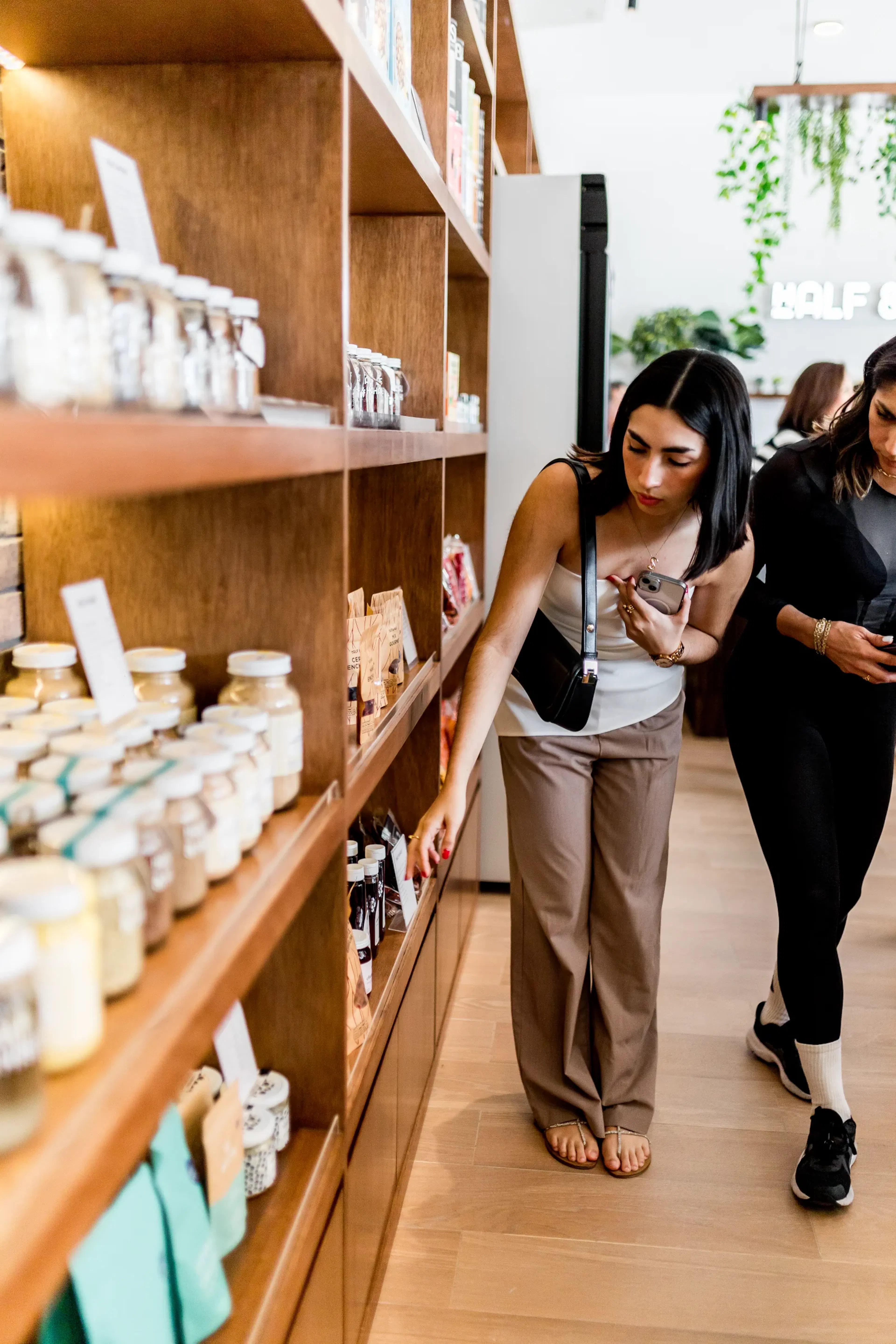 Una mujer de cabello largo y oscuro compra en una acogedora tienda con estantes de madera llenos de frascos. Se ve concentrada y comprometida, creando un ambiente cálido y acogedor.