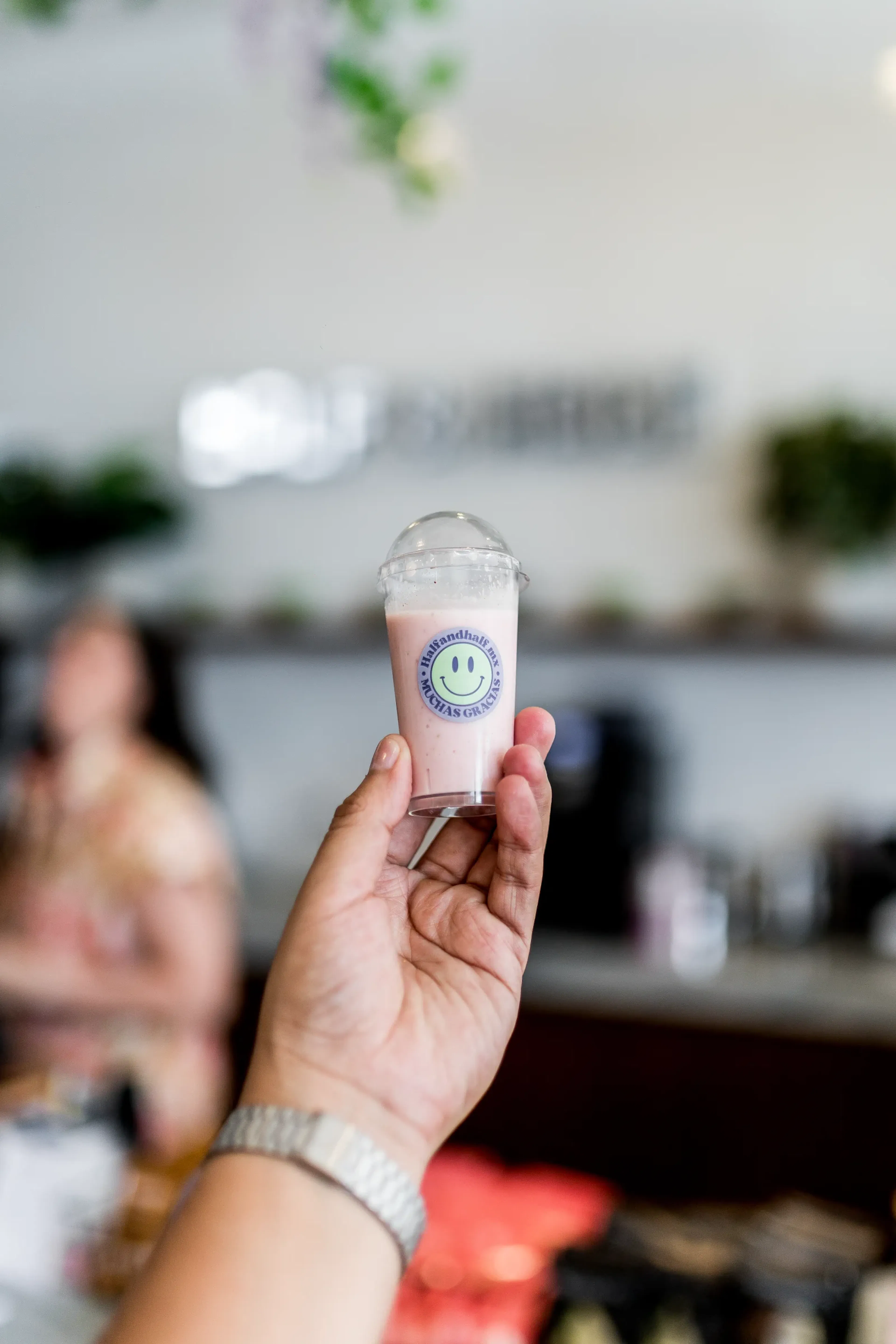 Una mano sostiene un vaso pequeño con un batido rosa, con el logo de una cara sonriente. El fondo es una cafetería moderna y desenfocada con un ambiente alegre.