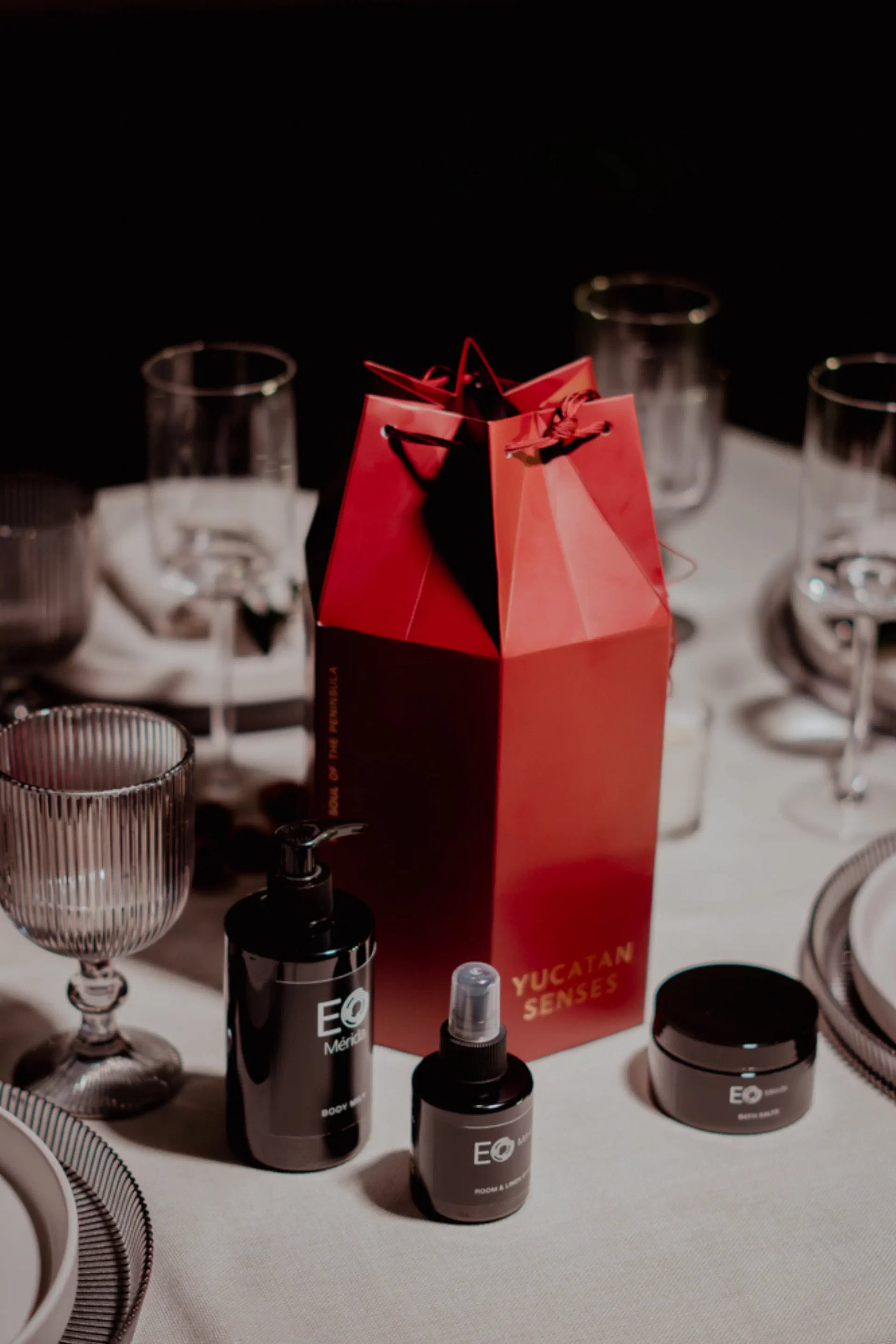 Bolsa de regalo roja con la etiqueta "Yucatan Senses" sobre una elegante mesa con diversos productos para el cuidado de la piel. La iluminación ambiental, la cristalería y los platos crean un ambiente lujoso y acogedor.