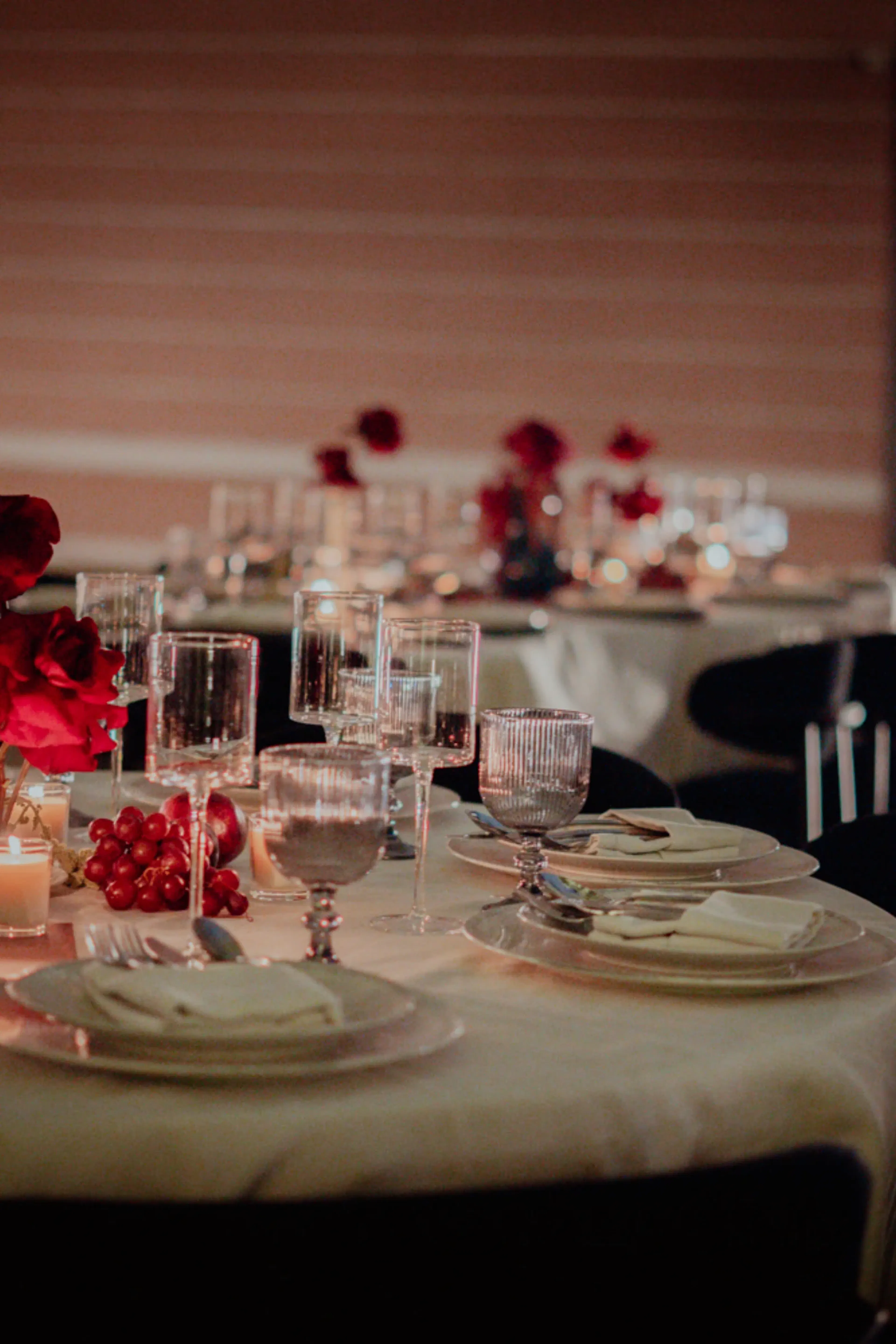 Una elegante mesa con copas de cristal, platos blancos y servilletas dobladas. Rosas rojas y velas aportan un ambiente romántico y cálido a la habitación con poca luz.